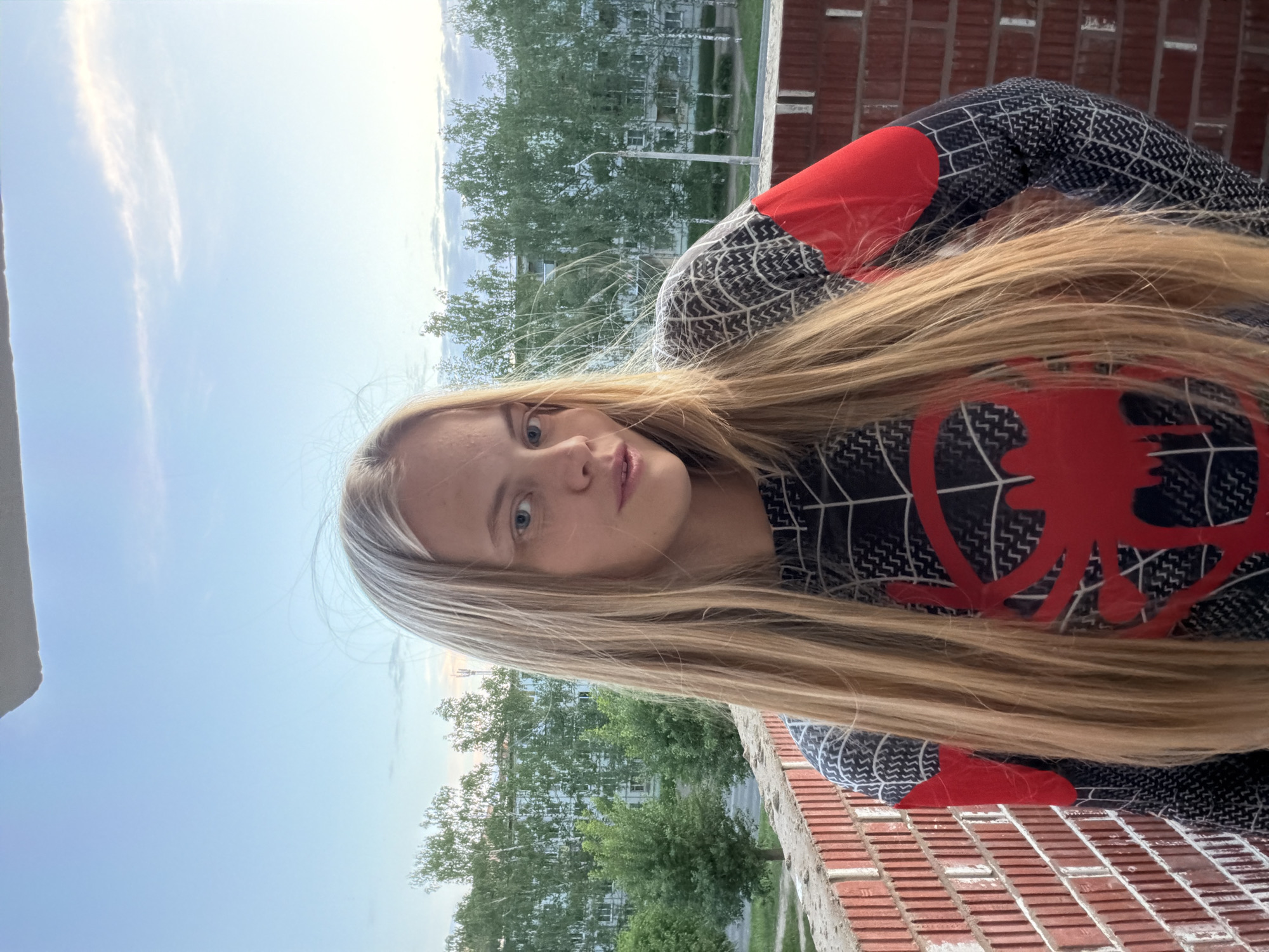 A young woman with long, straight blonde hair is posing for a photograph, likely a selfie, on what appears to be a balcony or ledge. She is wearing a Spider-Man themed outfit, with a prominent red spider emblem on the front. The background shows a cityscape with trees and buildings.
The photograph appears to have been taken from a high vantage point, as the view includes rooftops and the sky. The location may be Izhevsk, Russia, given the prompt. The scene suggests an outdoor, urban setting with natural elements like trees interspersed within the built environment. The image is composed in a way that highlights the subject and her outfit while providing context through the background scenery.