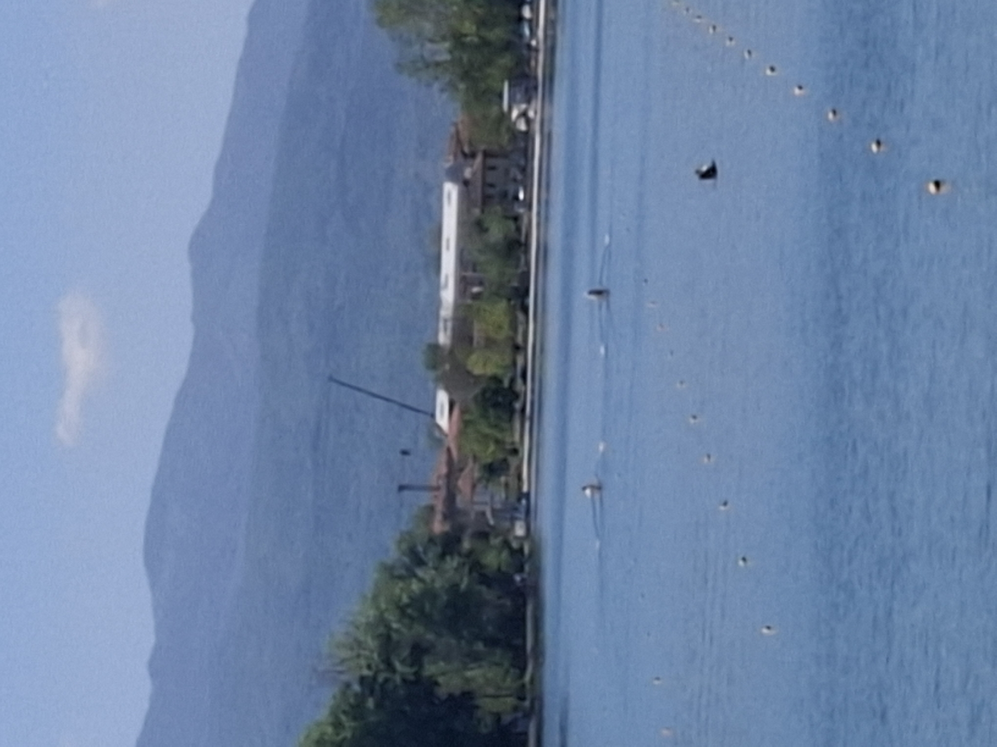 The image depicts a shoreline with mountains in the background. The shoreline features buildings and trees along the coast. A series of buoys is visible in the water. The scene appears to be in Plovdiv, Bulgaria, based on geographical characteristics and architecture.