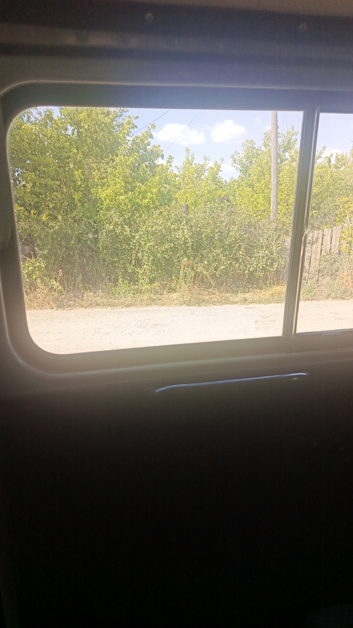 The image shows a view through a window looking out onto a rural scene. The foreground is dominated by the dark interior of a vehicle, with the bottom edge of the window frame visible. The window itself is rectangular with rounded corners. Outside the window, a dirt road or path runs horizontally across the frame. Beyond the road, a dense line of green foliage, likely trees and bushes, stretches across the middle ground. The foliage is lush and appears to be in full leaf, suggesting a summer or late spring setting. In the upper portion of the scene, the sky is visible, with a few white clouds scattered against a pale blue backdrop. To the right of the foliage, a wooden fence is partially visible, along with a utility pole. The overall impression is one of a peaceful, sunlit rural landscape seen from within a vehicle. The specific location in Zolotaya Step, Russia, is not visually identifiable from the image itself, but the scene depicts a typical rural environment consistent with such a location.