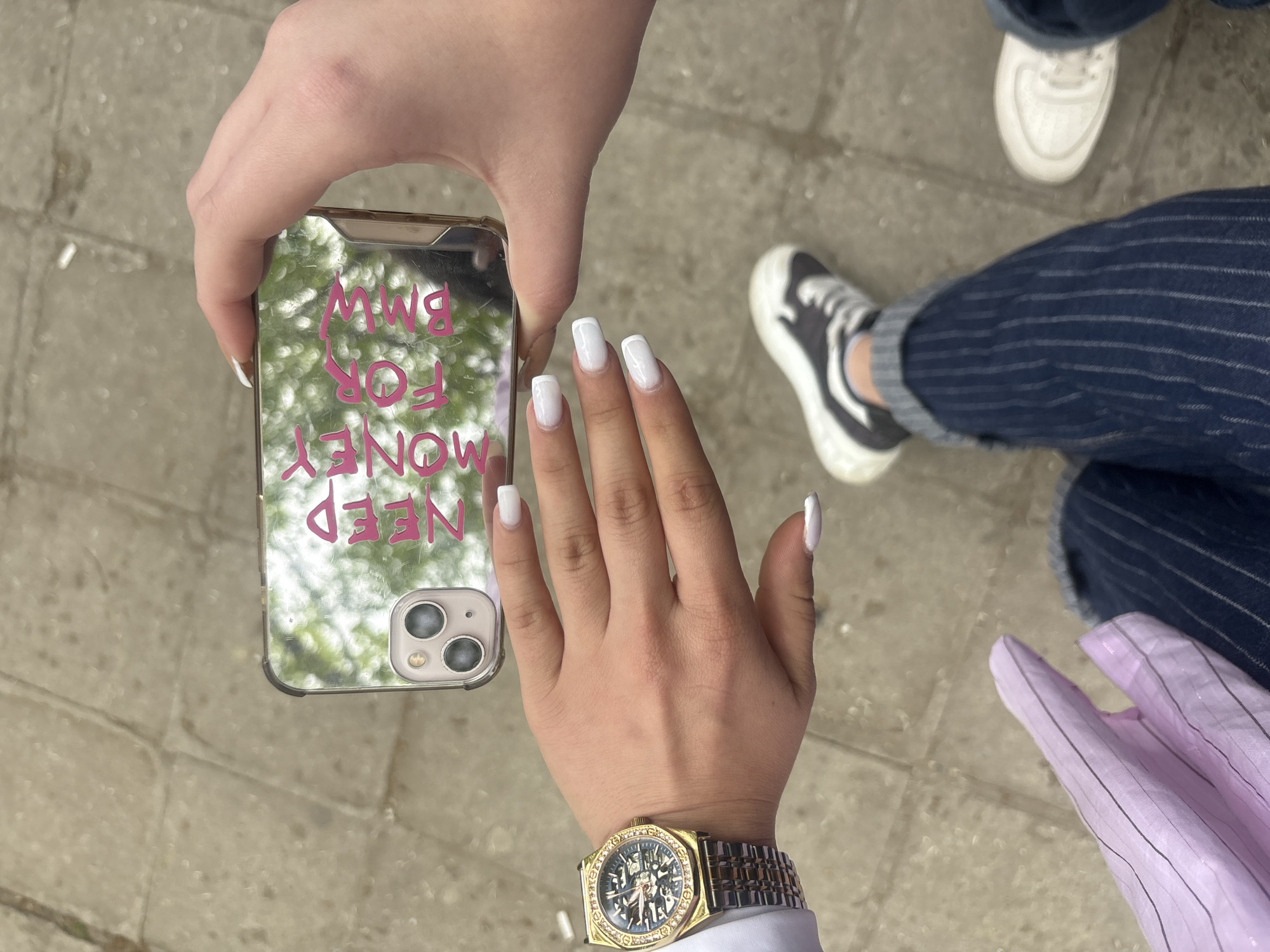 A view from above shows a person holding a smartphone with a reflective case bearing the pink text "NEED MONEY FOR BMW." The person has manicured white nails and wears a gold-colored watch. Beside this person are the legs of another person wearing pinstripe jeans and sneakers, one white and one black and white. A partially visible pink garment also appears in the scene. The setting is an outdoor public area paved with gray stones.