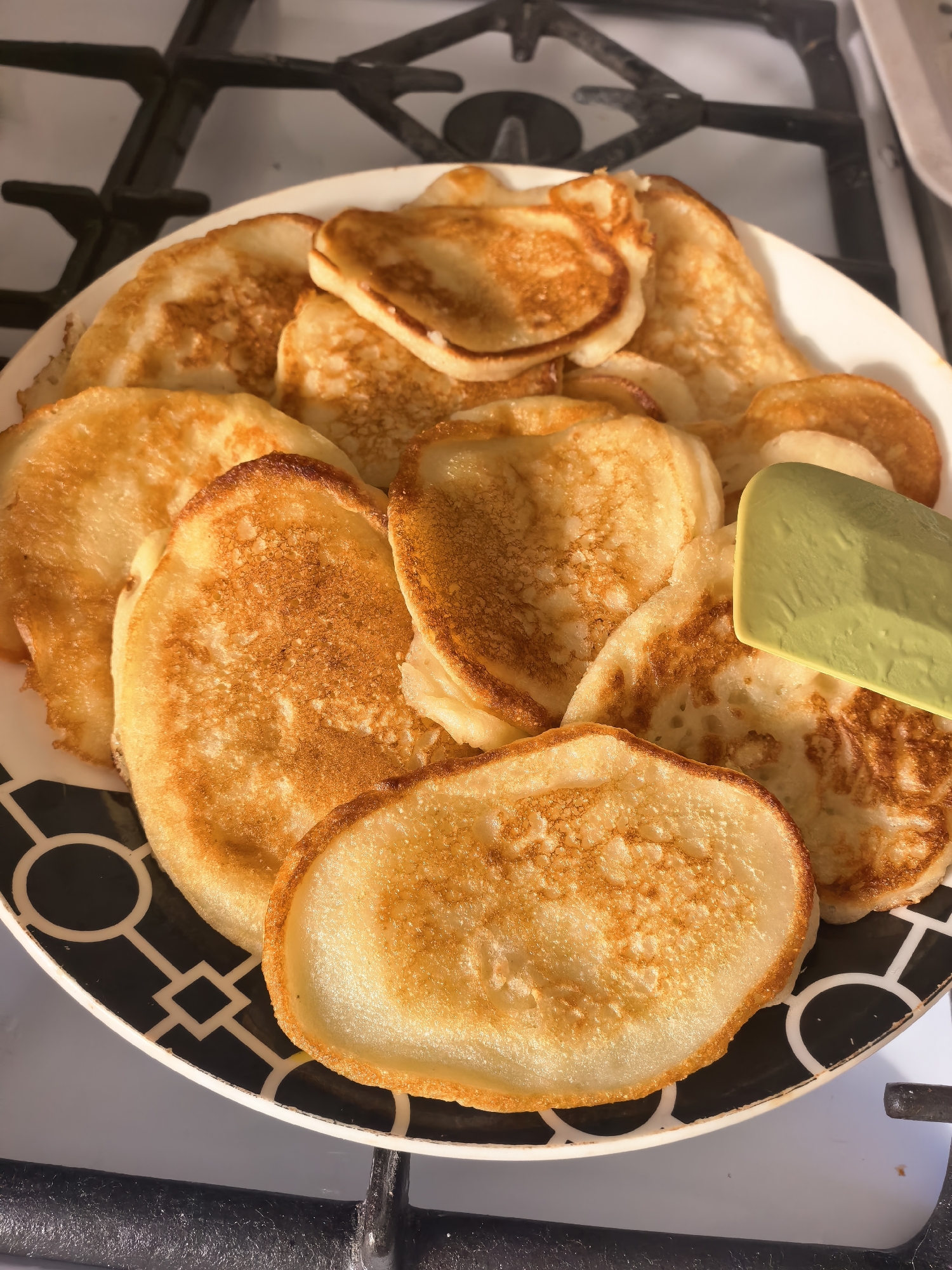A plate of pancakes sits on a gas stovetop. The plate is white with a black and white geometric pattern around the edge. The pancakes are golden brown, piled high on the plate, and a green spatula rests among them. The stovetop is black and metallic. There is no indication of Sterlitamak, Russia in the image.