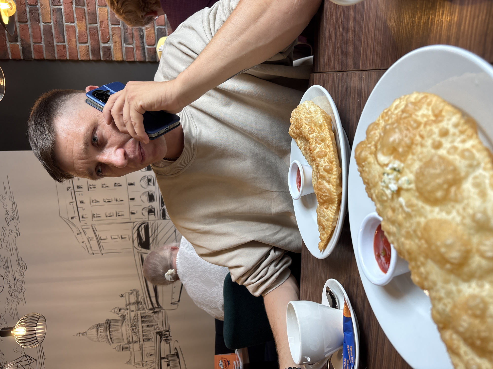 A man is sitting at a table in a restaurant, holding a blue smartphone to his left ear. He is wearing a beige sweater. On the table are two white plates, each with a fried pastry and a small white dish filled with red sauce. There is also a white cup and saucer. A drawing of a building resembling the Senate and Constitutional Court of Russia building is visible on the wall. The photograph was taken inside a restaurant in the city of Saint Petersburg, Russia.