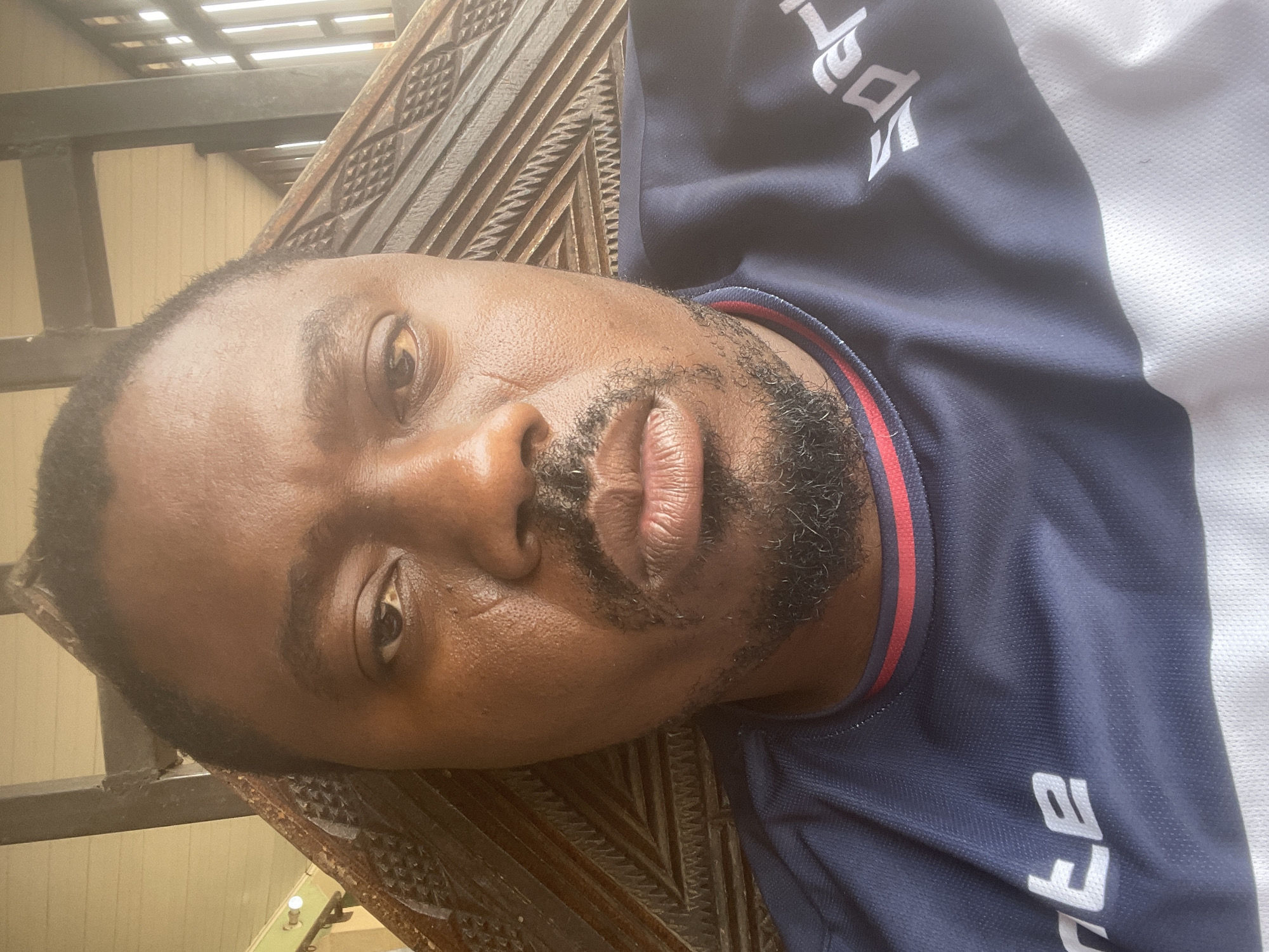 A close-up, rotated portrait features a man with dark skin and a short, neatly trimmed beard. He looks directly towards the camera with a neutral expression. He is wearing a blue athletic shirt with white sleeves and a red and white stripe around the collar. Partial white text, appearing to be "a pe" or similar characters, is visible on the blue section of his shirt.
The background shows a dark brown wooden surface with intricate, repetitive carved patterns, possibly part of a piece of furniture like a chair or headboard. Above and behind this wooden structure, metal bars are visible, along with a light-colored wall or building in the distance, and horizontal slats that could be part of an awning or roof. The bright, natural lighting suggests the photo was taken during the daytime, possibly in an outdoor shaded area or near a window. The setting is likely in Barakin Akawo, Nigeria, as provided by the location context.