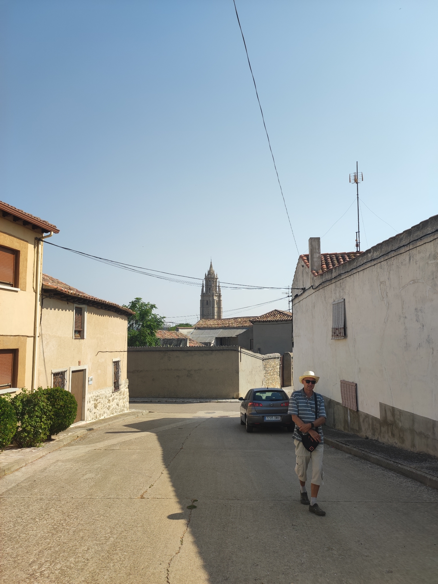 In Ampudia, Spain, a man wearing a straw hat and a striped shirt walks down a narrow, sun-drenched street. The man is carrying a bag across his chest. A dark grey car is parked on the left side of the street, and the street is lined with traditional Spanish houses. In the distance, a tall church spire rises above the rooftops, indicating the presence of the Church of San Miguel. The sky is clear and blue, with a few power lines crisscrossing the frame. The overall scene depicts a quiet, picturesque street in a Spanish town.