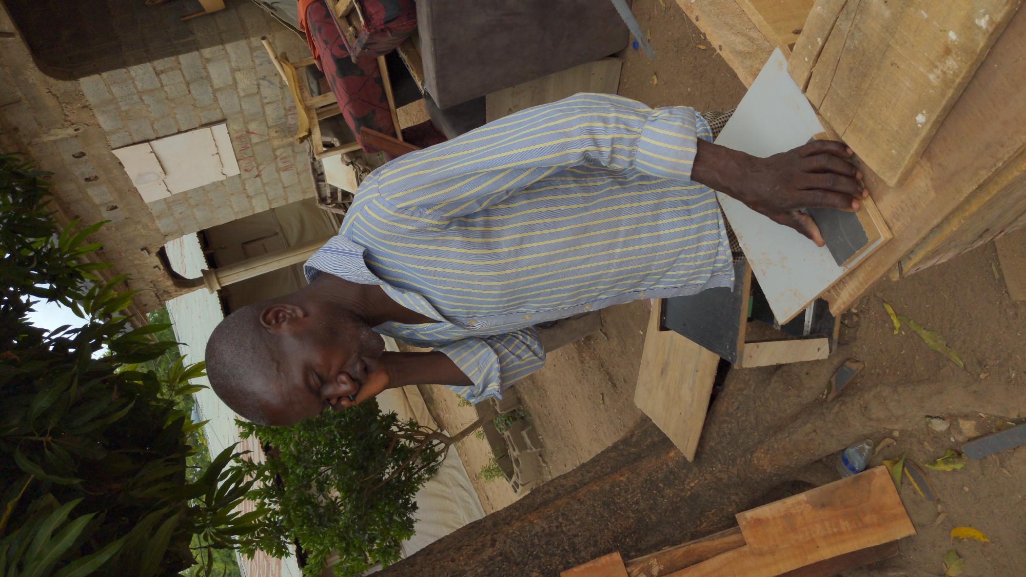 An outdoor scene in Masaka, Nigeria, depicts a man engaged in a manual task during bright daylight. The man, with dark skin and a short buzz cut, wears a light blue and yellow striped long-sleeved shirt. He is leaning over a wooden structure, carefully pressing a small, dark gray rectangular object against a white panel with his right hand. His expression appears focused or pensive, with his eyes partially closed as he concentrates on his work.
The setting is characterized by a dirt ground and a mix of construction materials and household items. Wooden planks and beams are prominent, forming a makeshift workbench or structure. In the background, there are concrete block walls of a building, possibly under construction or renovation. Red text, partially legible as "SLEE..." and "MEAS" or "MARS," is visible on one of the concrete blocks, suggesting construction markings. Scattered around are a plastic bottle, yellow leaves, and various pieces of furniture, including a brown sofa and another item with red patterned fabric. Green foliage from trees or bushes frames the left side of the image, adding a natural element to the otherwise functional space. The bright, even lighting indicates clear weather during the day.