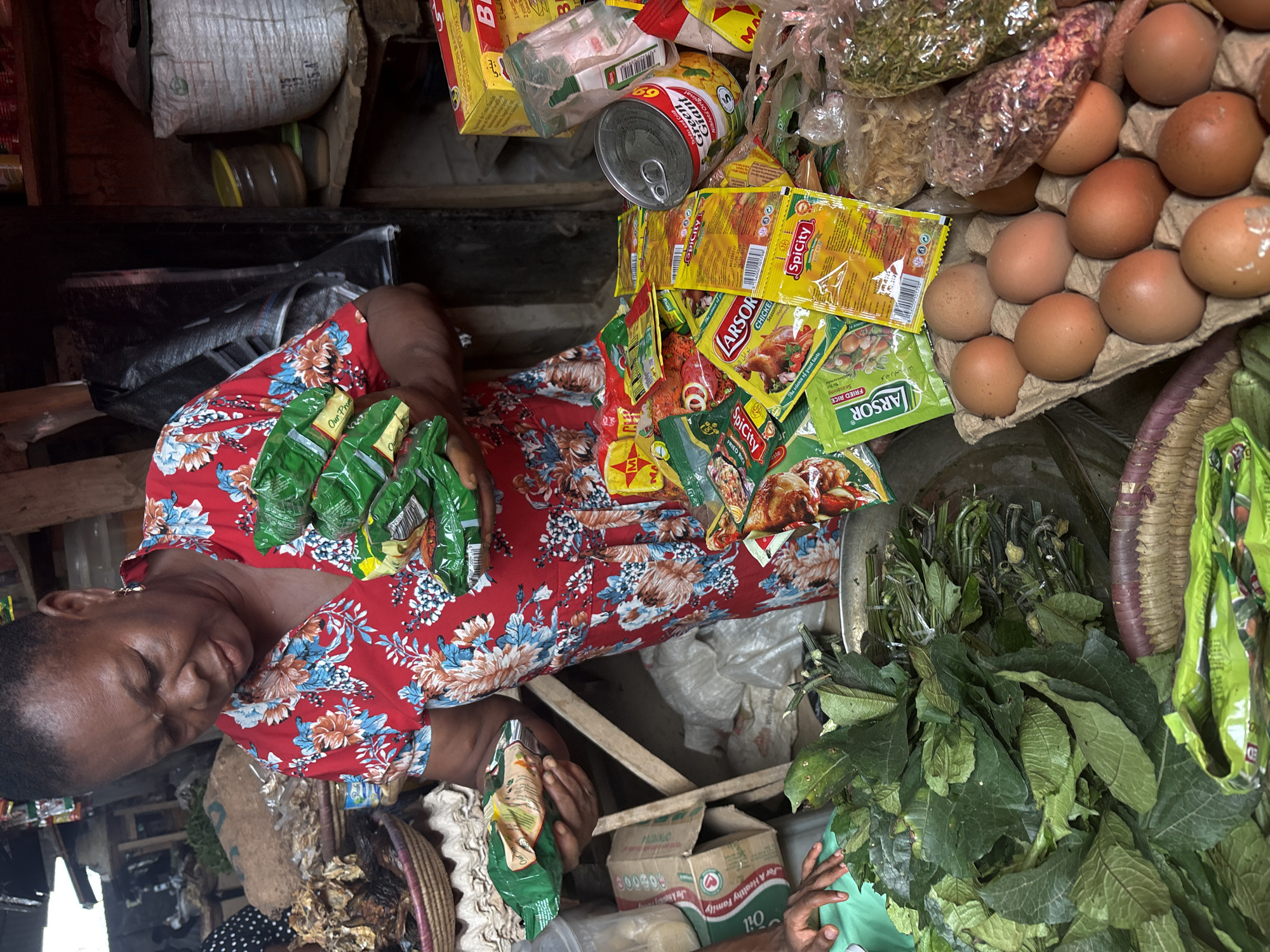 A woman in a red floral top sits in a stall in Iddo Tudunwada, Nigeria, surrounded by various grocery items. The stall features items such as eggs in a cardboard carton, leafy green vegetables, spice packets labeled "SpiCity" and "ARSOR," a "Green Giant" canned vegetable, bags of dried goods, and cooking oil. The woman is interacting with the items, and the scene is crowded with merchandise.
