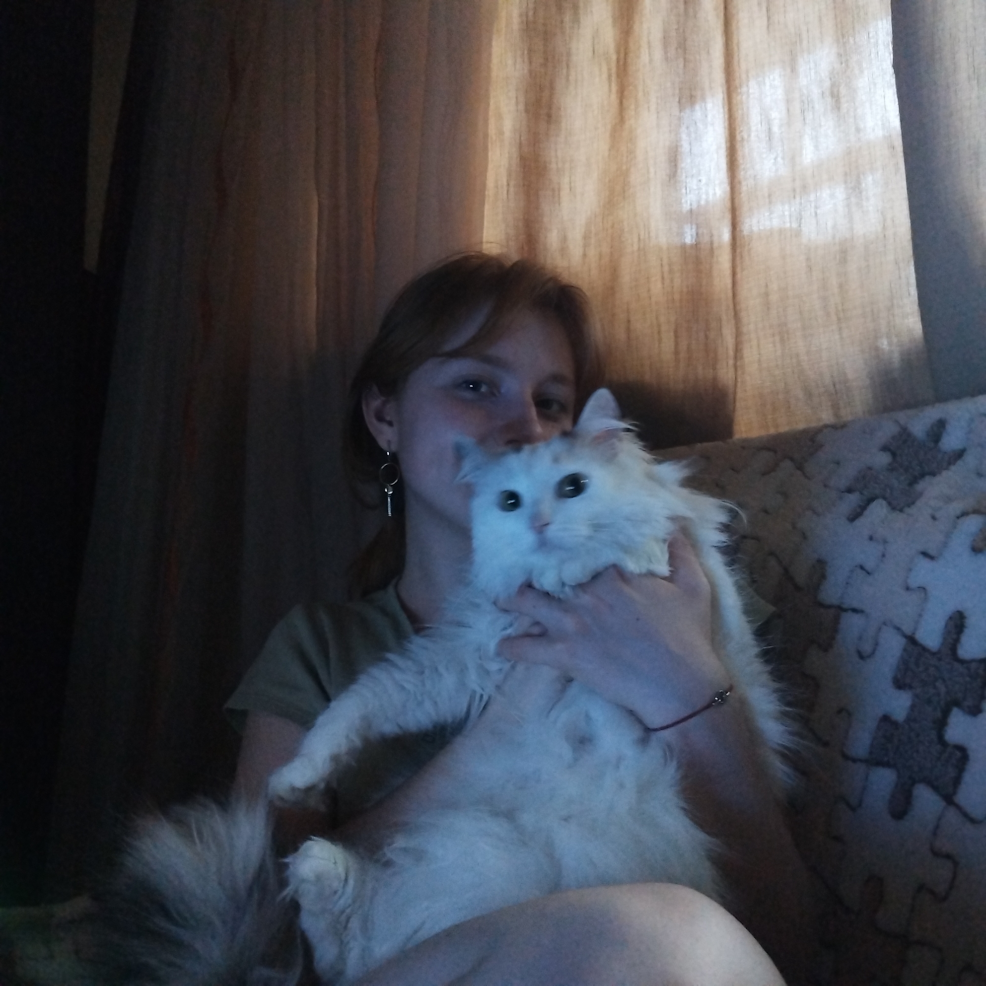 A person with short reddish-brown hair is holding a fluffy white cat. The person is wearing a green t-shirt and a silver earring. The cat has long white fur and green eyes. They are sitting on a couch with a puzzle-piece patterned covering. Behind them are draped curtains. The location and city of Yekaterinburg, Russia cannot be determined from the image.