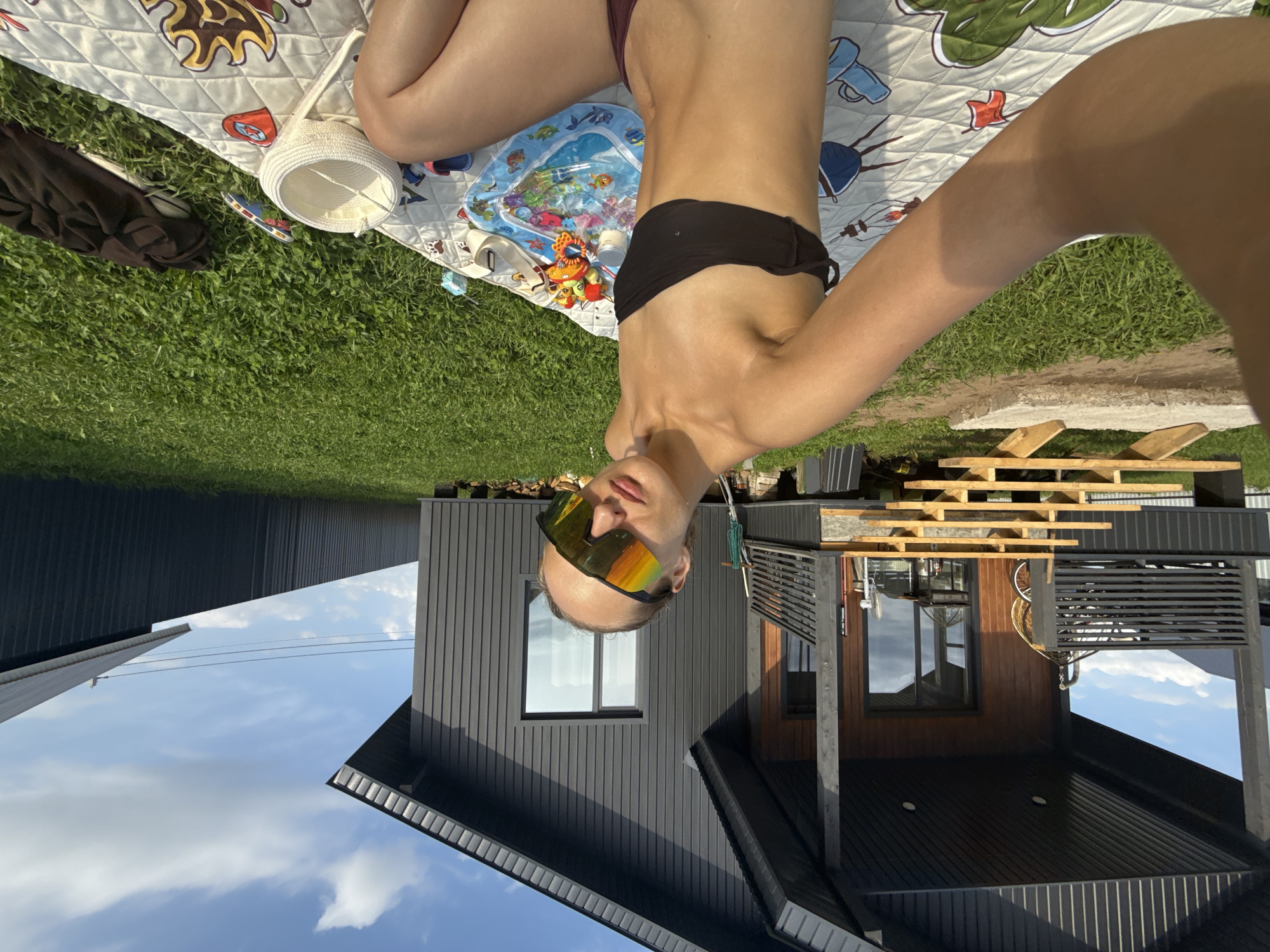 In the city of Podshivalovo, Russia, a young woman is taking a selfie outdoors on a sunny day. She is sitting on a checkered blanket spread on a grassy lawn in front of a modern, dark-sided house with a wooden porch and stairs. The woman is wearing a black bikini top and maroon bikini bottoms, along with large, reflective sunglasses. On the blanket next to her are various items, including a small inflatable pool with toys, a white woven basket, and a folded dark garment. The background shows the house and a fence, under a bright blue sky with white clouds.