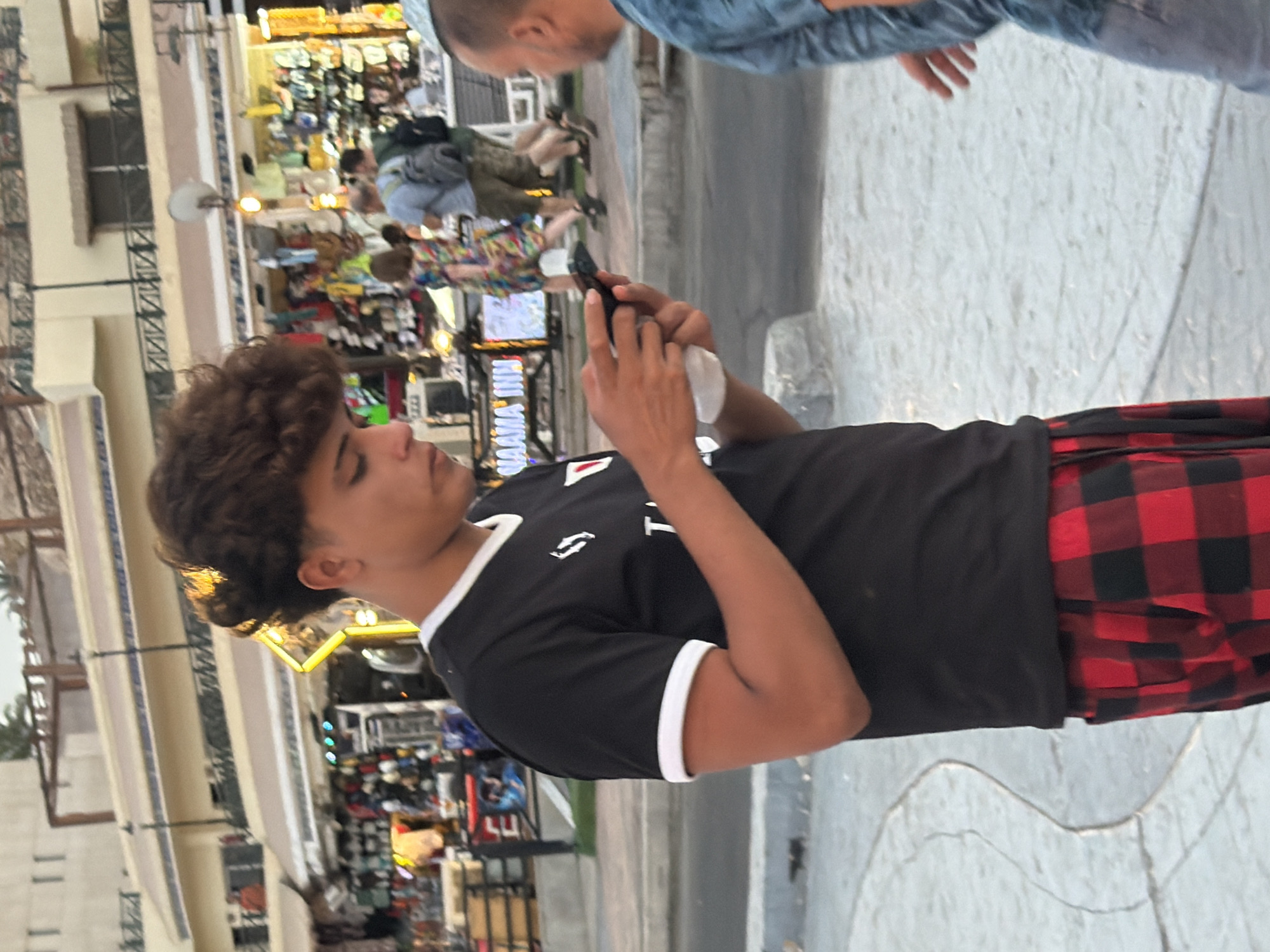 In Sharm El Sheikh, Egypt, a young man with curly brown hair stands outdoors, looking at his smartphone. He is wearing a black t-shirt with white trim on the collar and sleeves, and red and black plaid pants. His right hand holds the phone while his left hand is extended, possibly holding a white tissue. In the background, shops line the street, displaying various goods. Several other people are visible in the background, including a man in a blue t-shirt and another with a backpack. The environment appears to be a public area with a paved walkway.