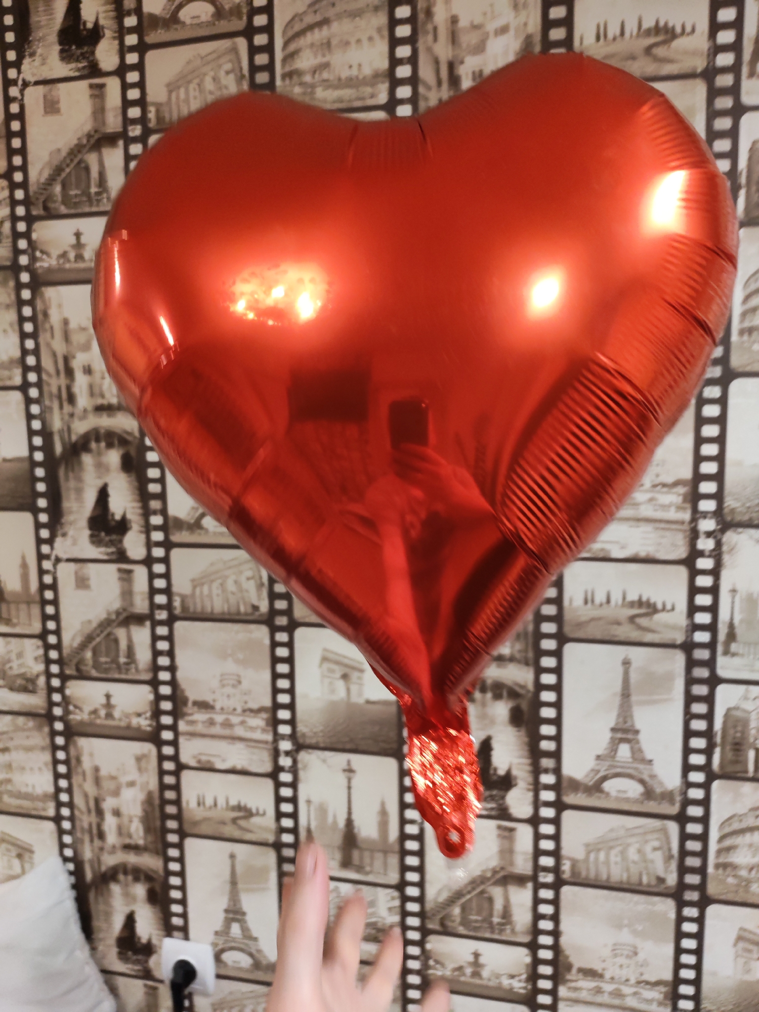 A red, heart-shaped balloon is the focal point of the image, appearing inflated and shiny. The background is a wallpaper with a design resembling strips of film, displaying images of various cities and landmarks, including the Eiffel Tower and the Colosseum. A hand is visible at the bottom of the frame, seemingly reaching towards the balloon. A white pillow and electrical outlet are at the bottom left of the frame. Based on additional knowledge, the image location is Chaikovsky, Russia, but it is not readily evident in the image itself.
