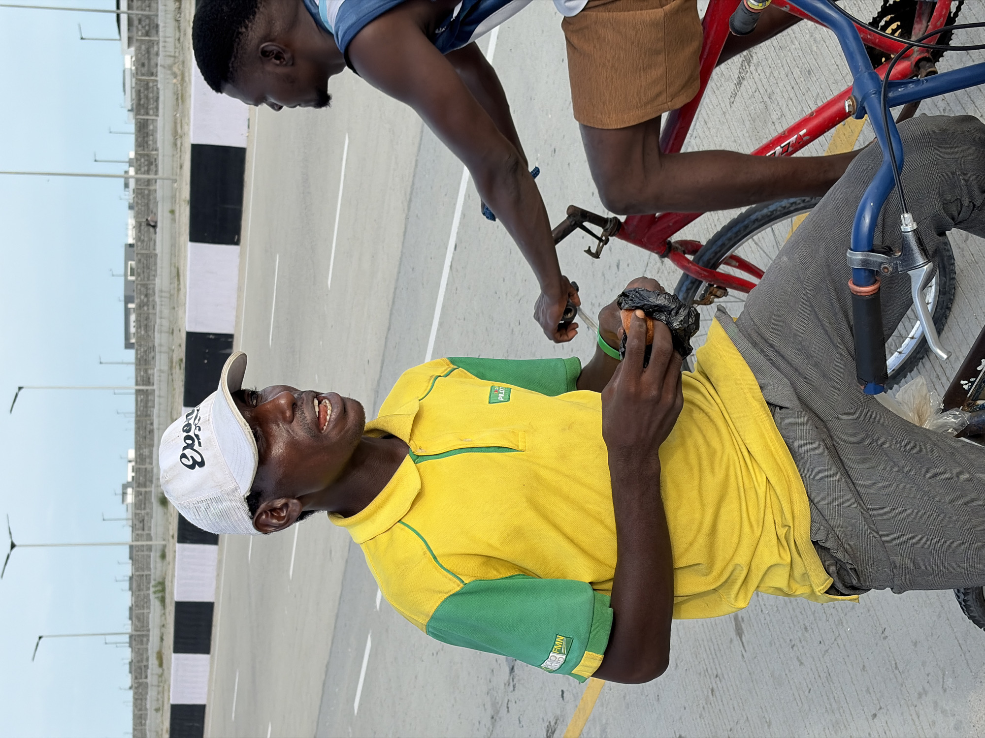 The image shows two men on bicycles on a paved road in Alaguntan, Nigeria. The man closest to the camera is wearing a yellow and green polo shirt, grey trousers, and a white baseball cap. He is holding a wrapped object in his hands. The second man is wearing a blue and white shirt and tan shorts. He is sitting on a red bicycle. The road is marked with white and yellow lines and bordered by a barrier with alternating black and white sections. Streetlights line the roadside. Buildings are visible in the background. The sky is overcast.