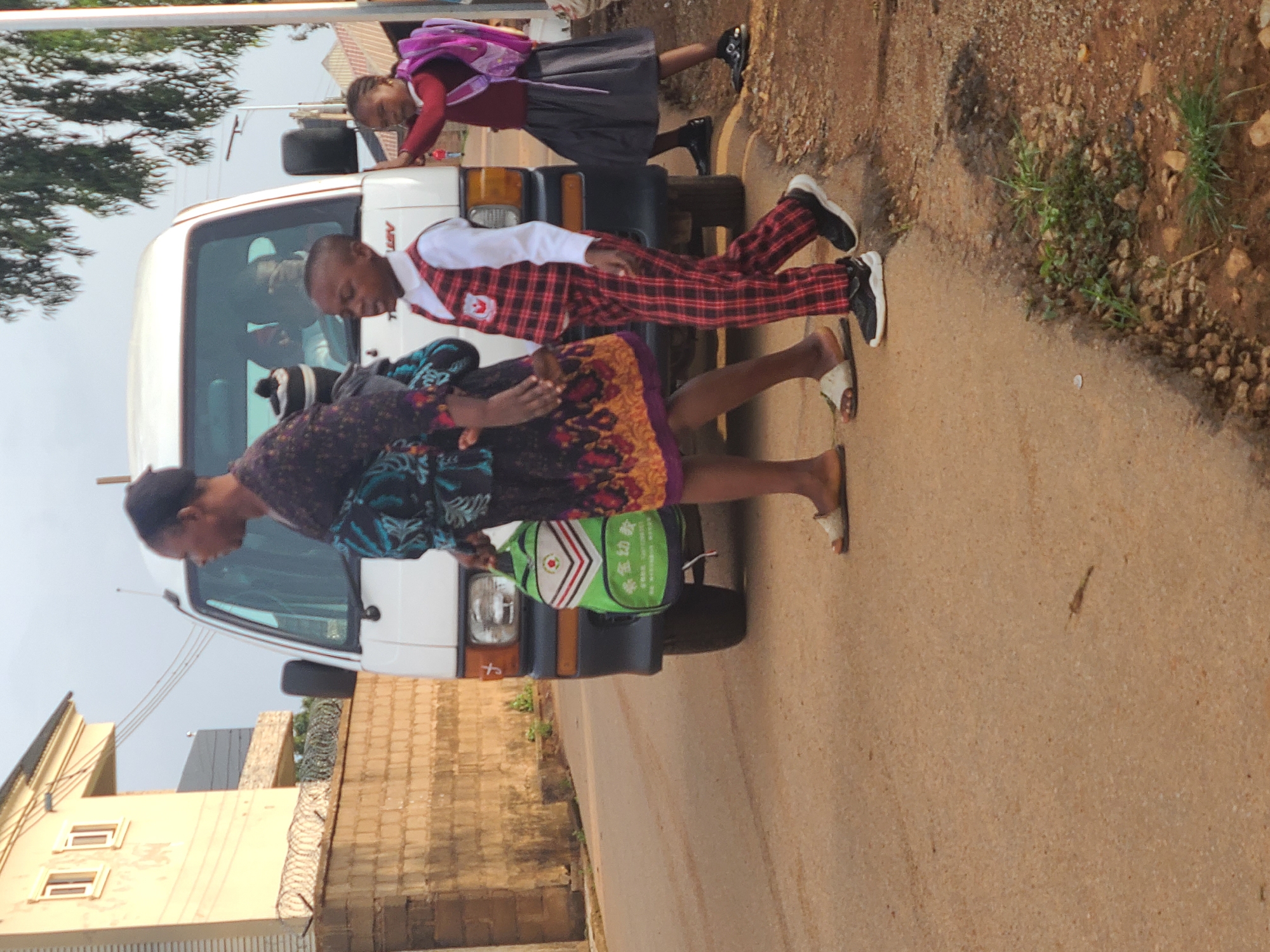 The scene shows a white van parked on the side of a paved road in Jos, Nigeria. Three people are present. An adult female wearing a patterned dress and sandals stands beside the front of the van, holding a green bag. Two children in school uniforms are near the van. One child in a plaid uniform stands facing the van while the other child, wearing a maroon uniform with a backpack, is facing away from the van with their hands on the hood. Buildings with fences and vegetation are visible along the roadside.