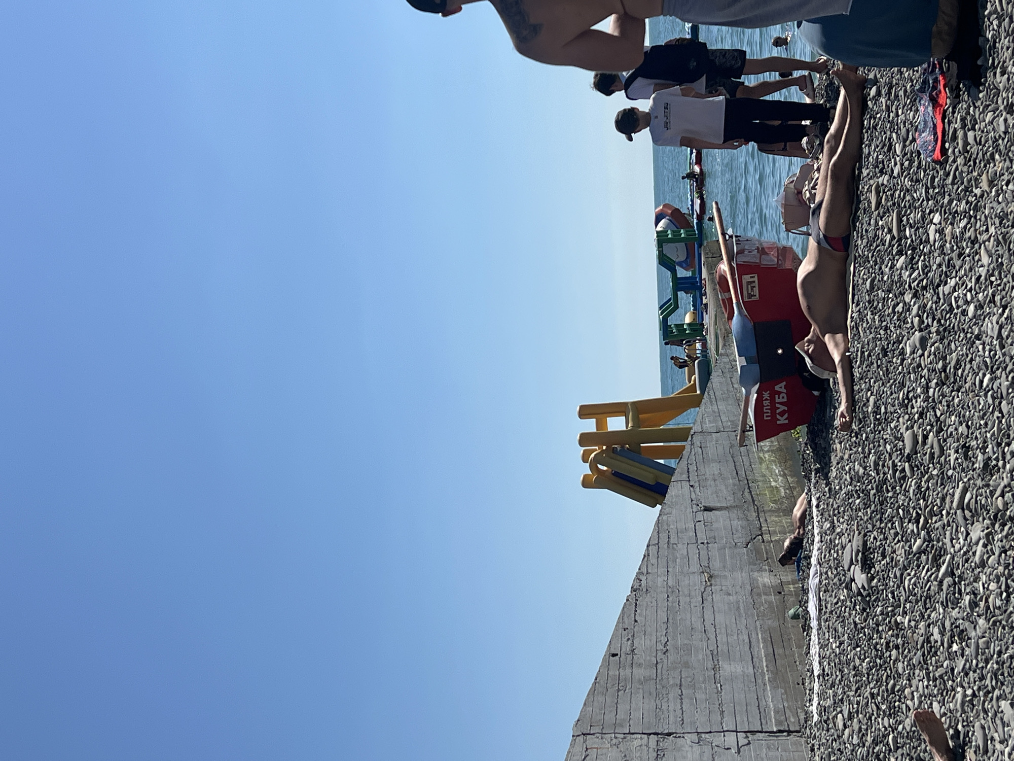 The image depicts a lively scene on a pebbled beach in Sochi, Russia, under a clear blue sky. In the foreground, a man is sunbathing on a towel, wearing swim trunks. To his right, another person's legs and lower torso are visible, also appearing to be relaxing on the beach. Further back, a group of young men stand near the water's edge.
Dominating the background is the Black Sea, with several inflatable water attractions, including slides and a large inflatable structure, visible in the water. A red lifebuoy with the inscription "ПЛЯЖ КУБА" (Beach Kuba) is placed on the shore. A concrete structure, possibly part of a seawall or promenade, runs along the left side of the image. The beach is covered with numerous grey and dark pebbles. The overall atmosphere is that of a sunny day at a popular tourist destination.