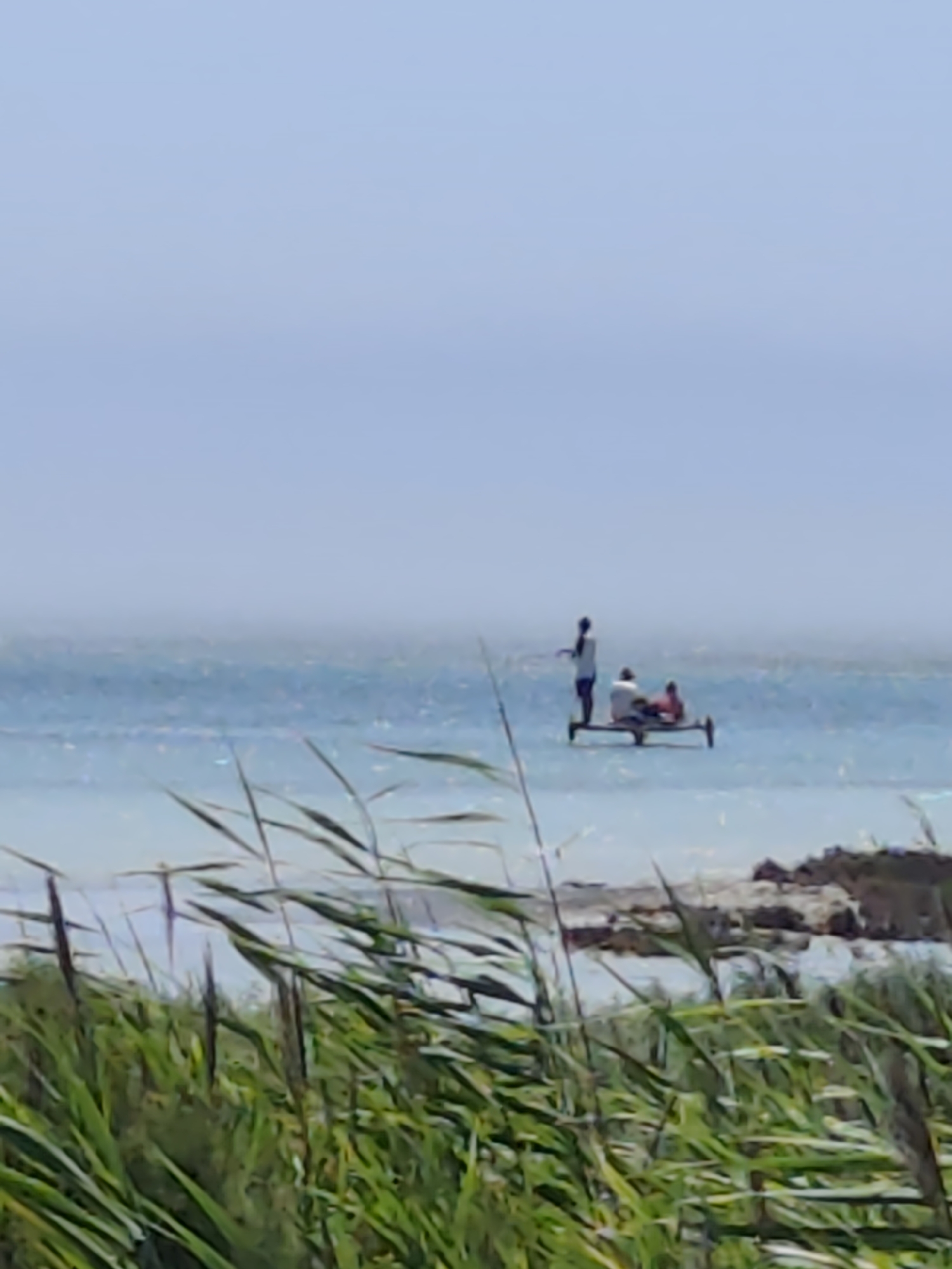 In the city of Aktau, Kazakhstan, a person stands on a small boat or raft, seemingly fishing. Two other individuals are seated on the same craft. The scene is set on a body of water, likely the Caspian Sea, with a hazy sky. Reeds and tall grass are in the foreground, obscuring the immediate shoreline and parts of the water's edge. The overall atmosphere is calm, with the water appearing somewhat choppy and the sky a pale, washed-out blue.