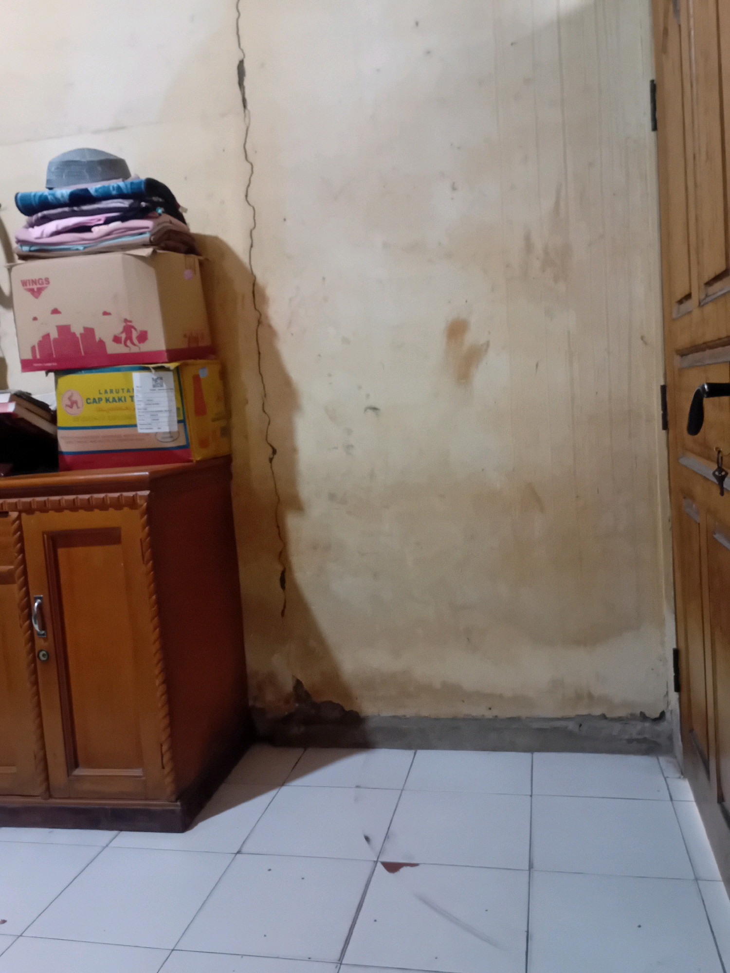 The image depicts an interior scene, likely a room in a residential or storage area in Pekanbaru, Indonesia. A wooden cabinet with a dark brown trim is visible on the left side of the frame. On top of the cabinet, there are several stacked cardboard boxes, one with the "WINGS" logo and a graphic of buildings and a person, and another box with Indonesian text including "LARUTAN CAP KAKI T". A pile of folded clothing, topped with a dark-colored prayer cap, rests on the boxes. The wall behind the objects is a light beige color, exhibiting signs of wear and tear, including a prominent vertical crack. To the right, a wooden door with vertical panels and a black handle is partially visible. The floor is covered with light-colored square tiles. The overall impression is one of a functional, albeit somewhat cluttered, storage space.