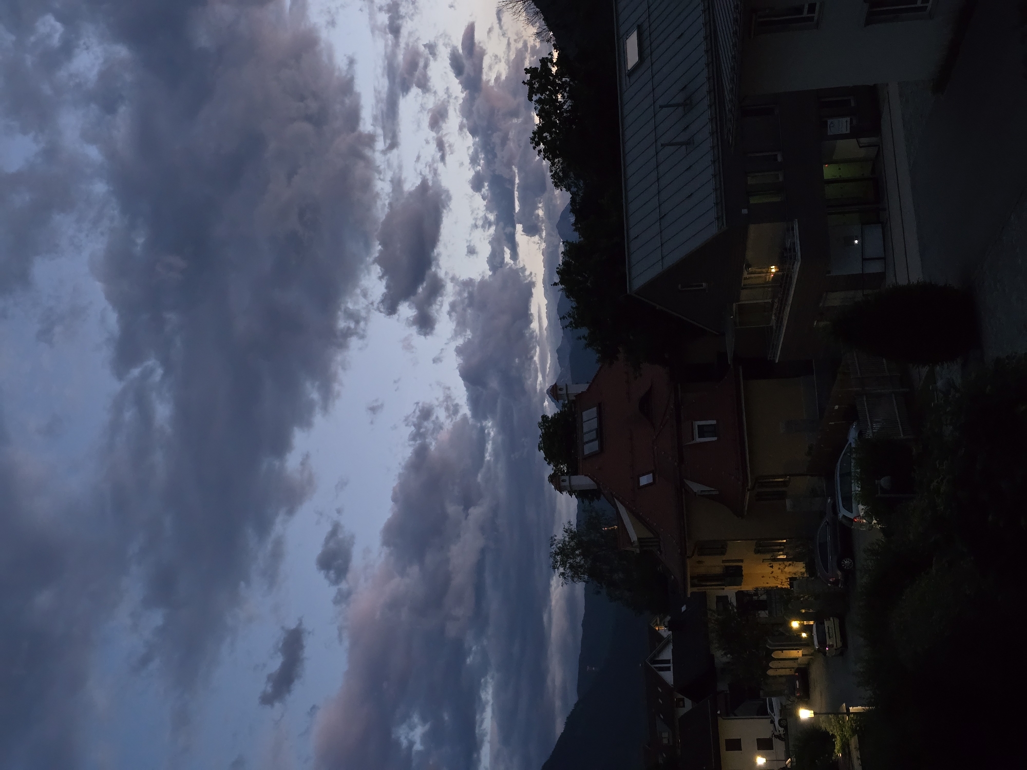 The image depicts a twilight scene in Garmisch-Partenkirchen, Germany, with a sky filled with dramatic clouds. In the foreground, the roofs of several buildings are visible, including a house with a red tiled roof and chimneys, and a modern building with a dark, sloped metal roof. Streetlights illuminate a street with parked cars, including a white hatchback and two SUVs. The background shows a densely wooded hillside and distant mountains shrouded in shadow. The overall impression is a calm, suburban evening in a mountainous region.
