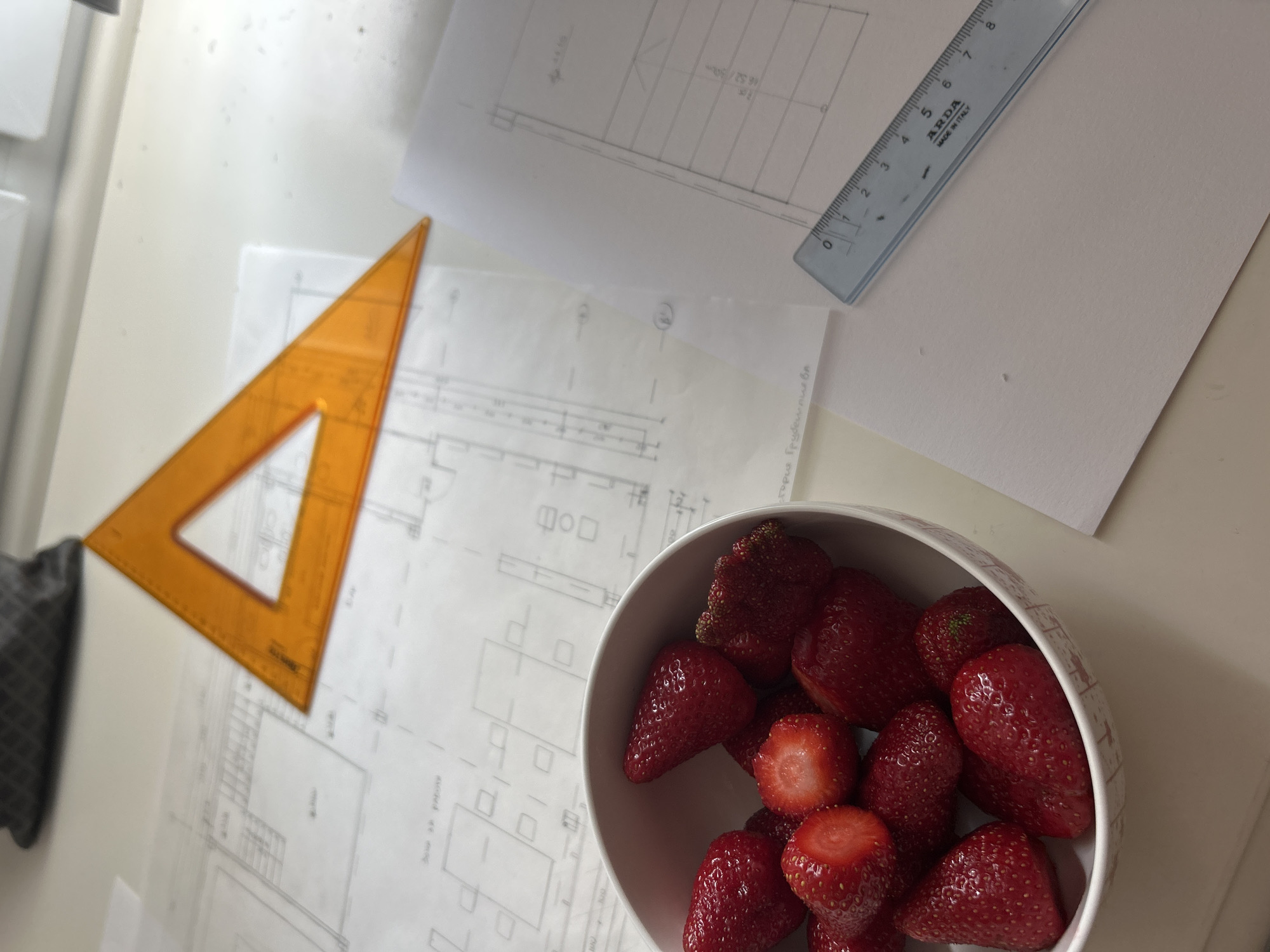 The image shows a desk with architectural drawings, a clear plastic ruler, a transparent orange triangle, and a bowl of strawberries. The setting is likely indoors, given the controlled lighting and presence of office supplies. There is no evidence indicating this is in Plovdiv, Bulgaria.