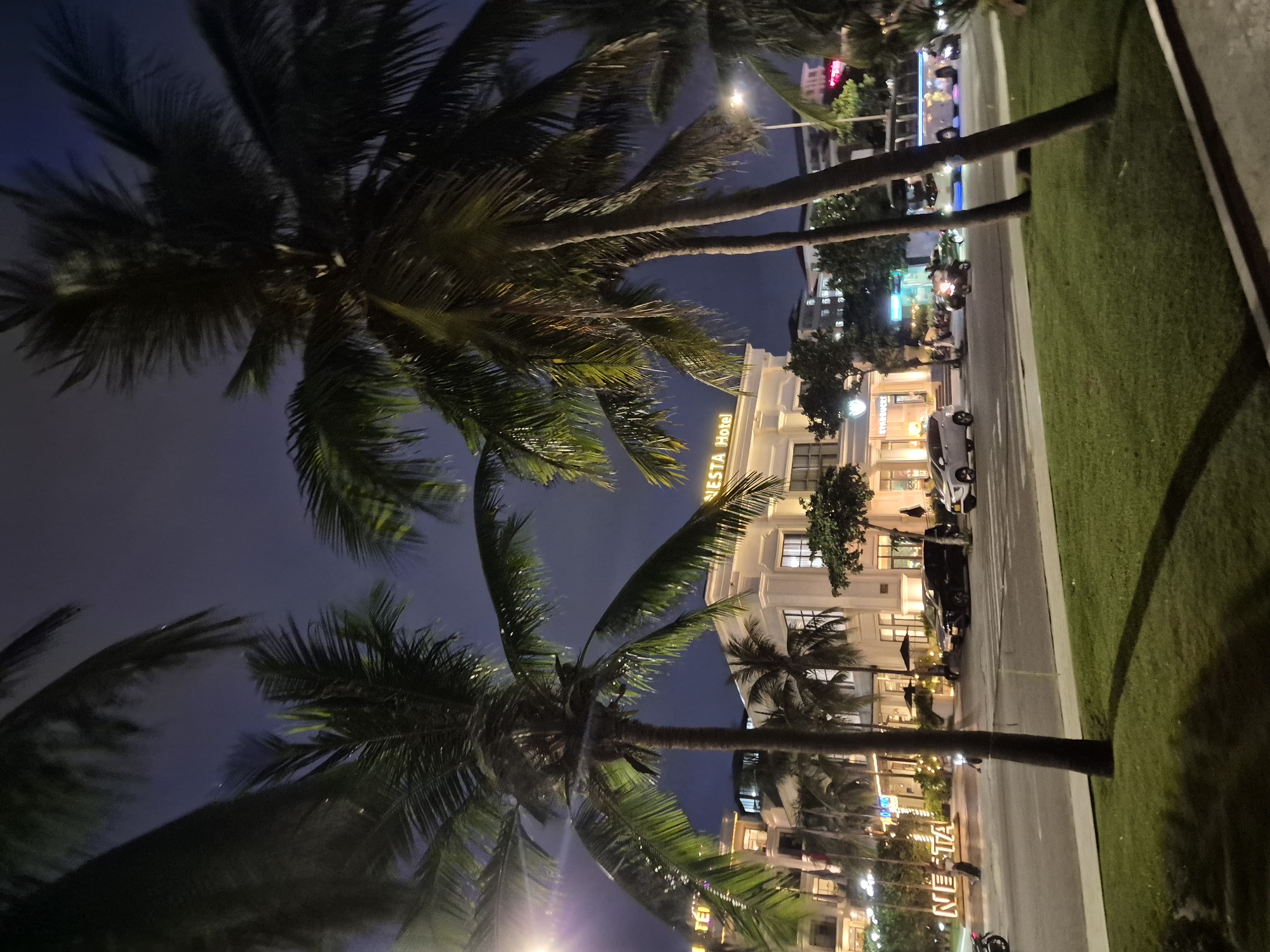 At night in Da Nang, Vietnam, a street scene unfolds with palm trees framing a building labeled "NESTA Hotel". The hotel's facade is illuminated, and its name is prominently displayed in a large, lit sign. To the left of the hotel, another lit sign for "NESTA" is visible. A Starbucks is also visible in front of the hotel. The street in front of the hotel is busy with traffic, including cars, motorcycles, and a bus. The ground is covered in green grass, and the sky is dark.