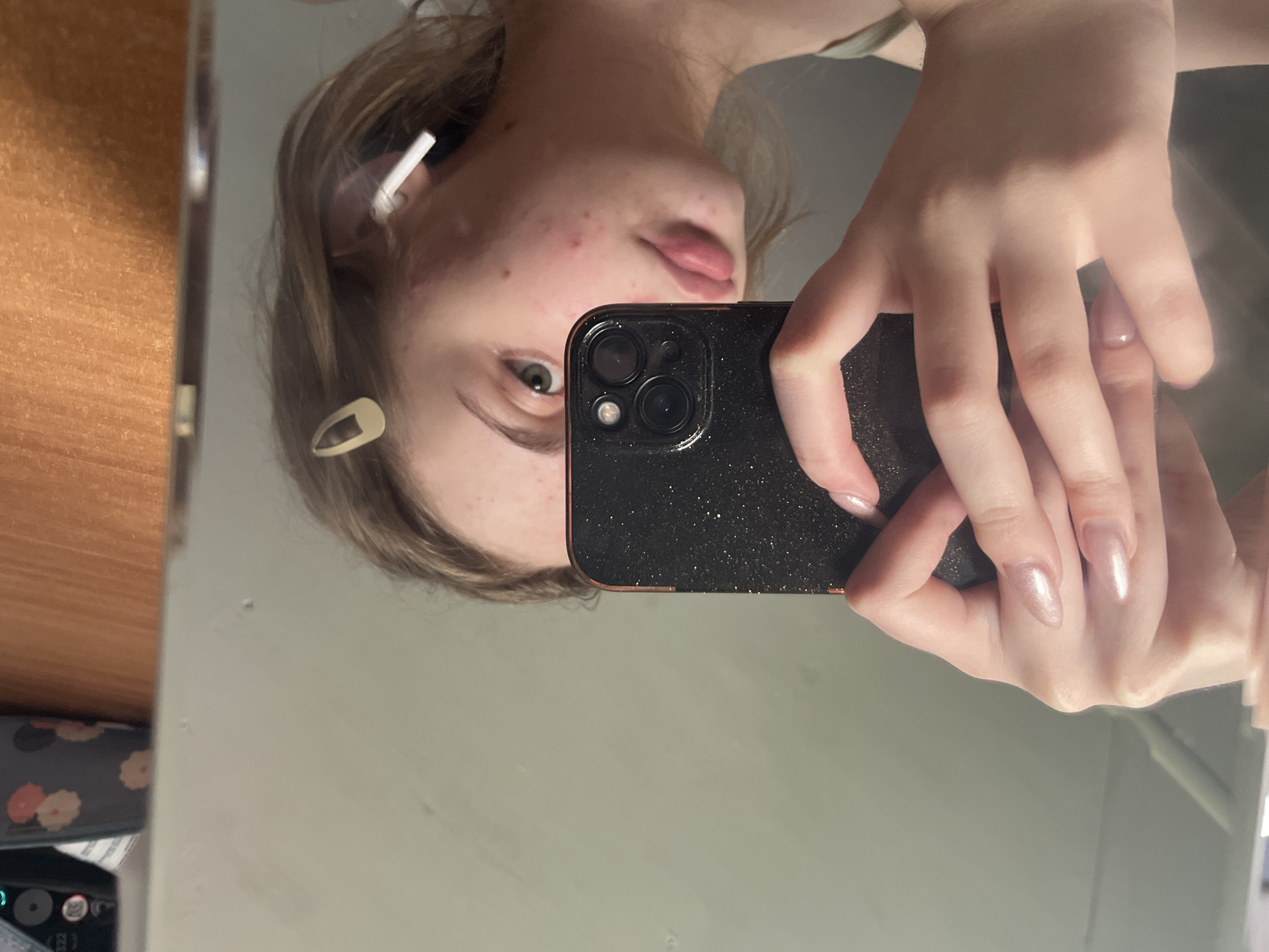 A young person with light skin and reddish-blonde hair is taking a selfie in a mirror. They are holding a black smartphone with a glittery case in their right hand, with their left hand visible, showing long, light-pink acrylic nails. The person is wearing a light-colored top and a white wireless earbud in their right ear. A gold-colored hair clip is visible in their hair. Their face has some acne blemishes. The background is a plain, light-colored wall and a wooden surface on the right. No specific location in Ashkadar, Russia can be determined from the image.