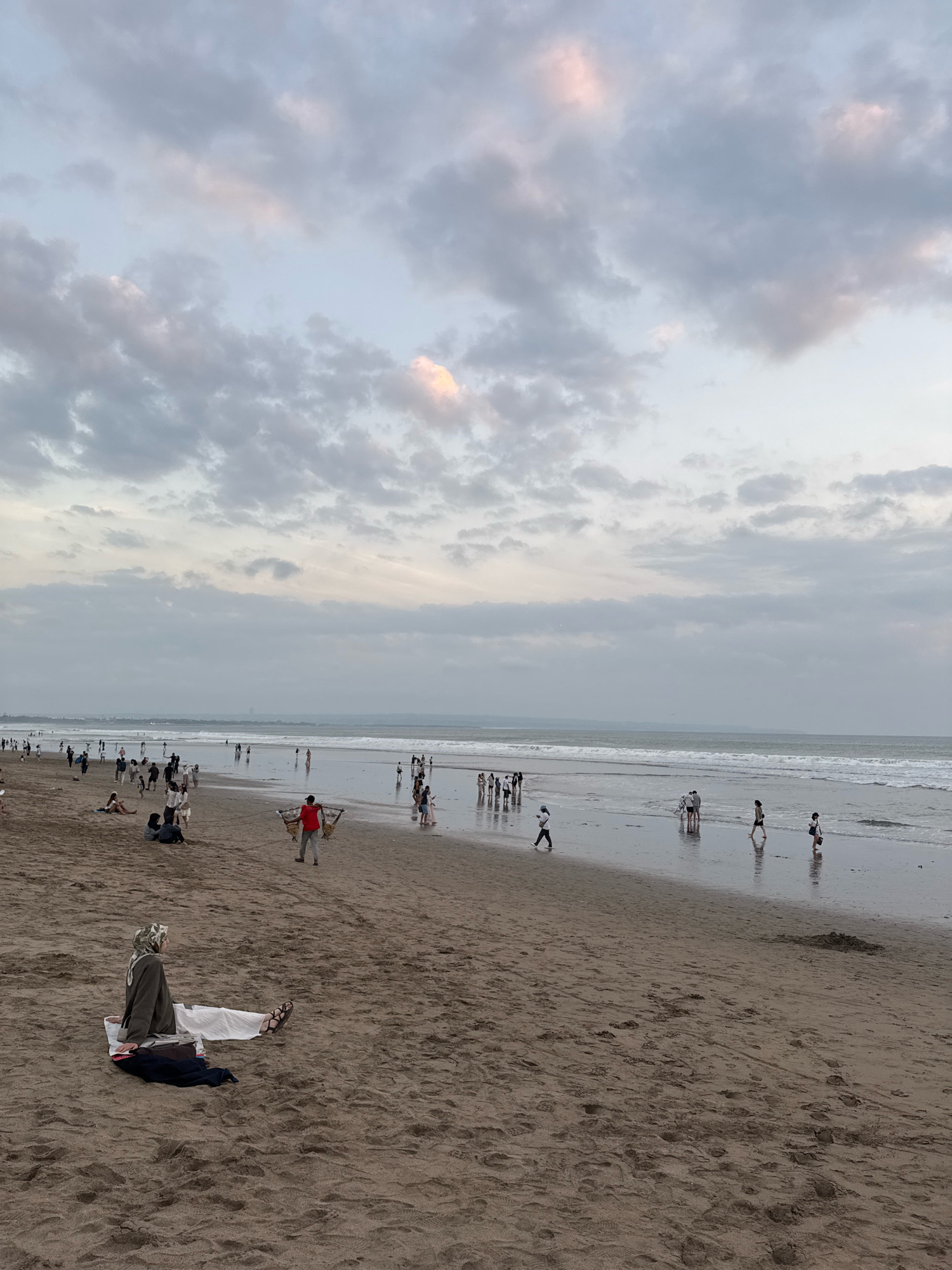 In Kuta, Indonesia, a wide, sandy beach stretches towards the ocean under a cloudy sky. Numerous people are scattered across the sand and the shallow water. In the foreground, a woman wearing a hijab and white pants sits on the sand, facing away from the viewer. Further down the beach, groups of people walk along the shoreline and in the water, with some standing and observing the ocean. A man in a red shirt is seen walking with a kite. The waves are moderate, and the overall atmosphere is of a public gathering on a beach.