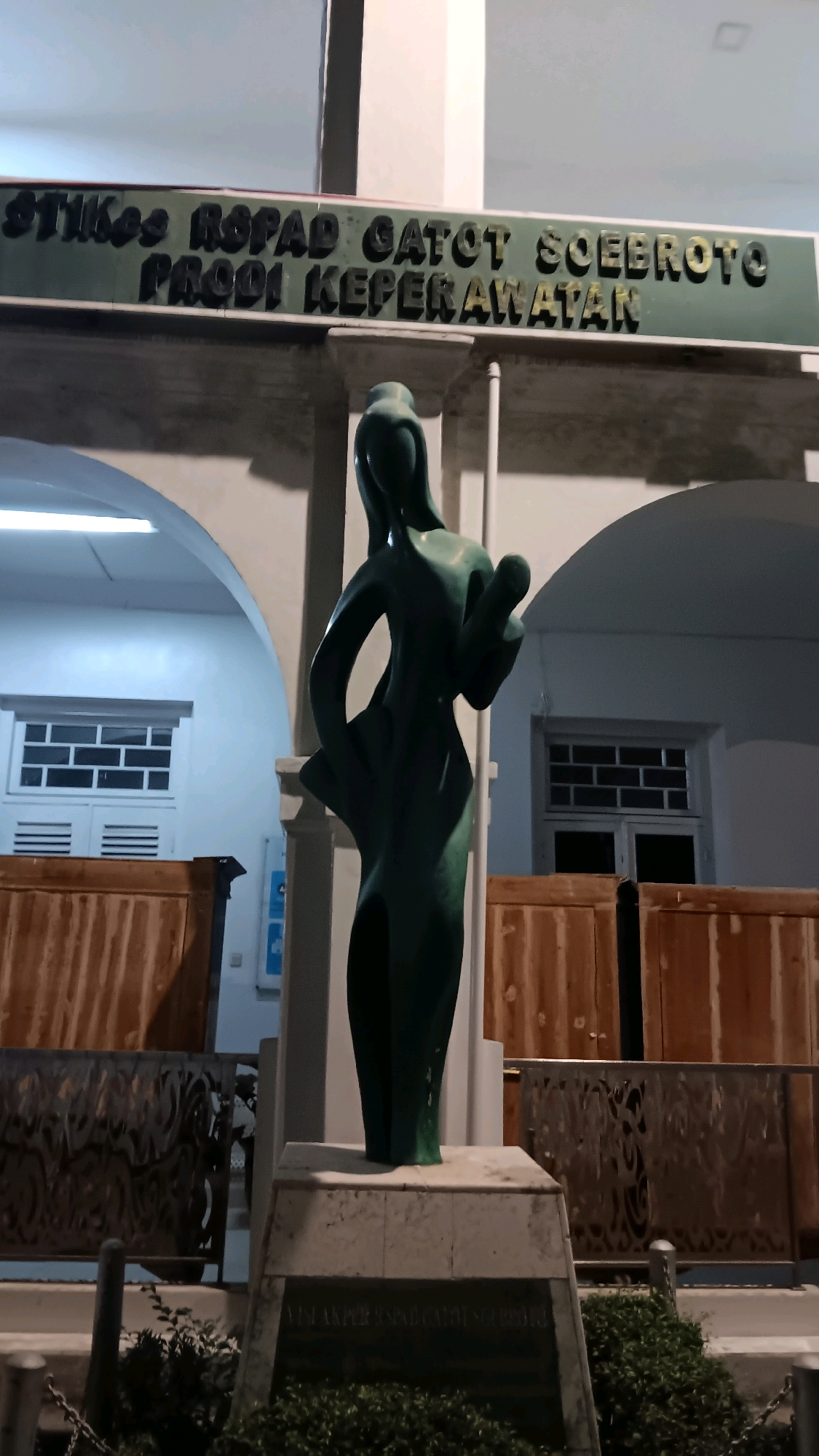 An outdoor, low-angle medium shot captures a dark green abstract statue of a woman holding a baby in front of a building at night. The statue stands on a stone pedestal, which has a plaque at its base. The building behind the statue has arched entrances, wooden paneling, and windows with horizontal slats. A green sign with dark, raised lettering is visible above the statue, reading "STIKES RSUP GATOT SOEBROTO PRODI KEPERAWATAN." The area is lit by artificial light, casting shadows and highlighting the green hue of the statue.