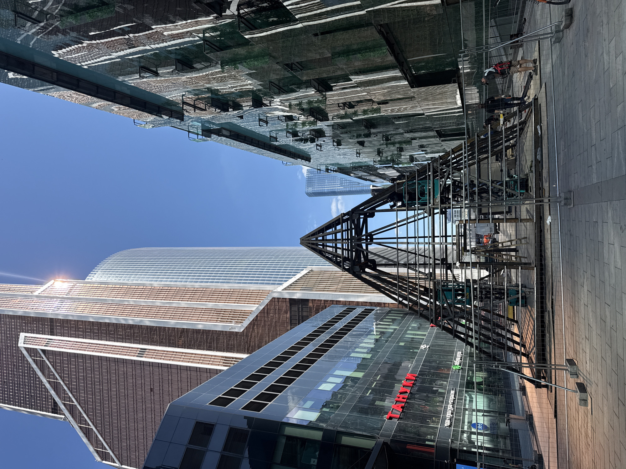 The image depicts an outdoor urban scene on a sunny day, likely in Moscow, Russia, given the architectural style of the skyscrapers. In the foreground, a temporary metal fence encloses a construction area. Inside the fenced zone, a large, pyramid-shaped metal framework is being erected. Two individuals are visible near the framework; one appears to be operating a scissor lift, while another stands on the ground observing the work.
In the midground, a modern glass-fronted building with red lettering that reads "ТАНУКИ" is situated to the left. Adjacent to this building, and to the right of the metal framework, is another building covered in green netting and scaffolding, suggesting ongoing construction or renovation.
The background features several imposing skyscrapers, characteristic of a modern business district. The tallest skyscraper, with its distinct brown and white facade, is particularly prominent. Another skyscraper with a predominantly glass exterior is visible further in the distance. The sky is clear and blue, indicating good weather. The ground is paved with rectangular bricks.