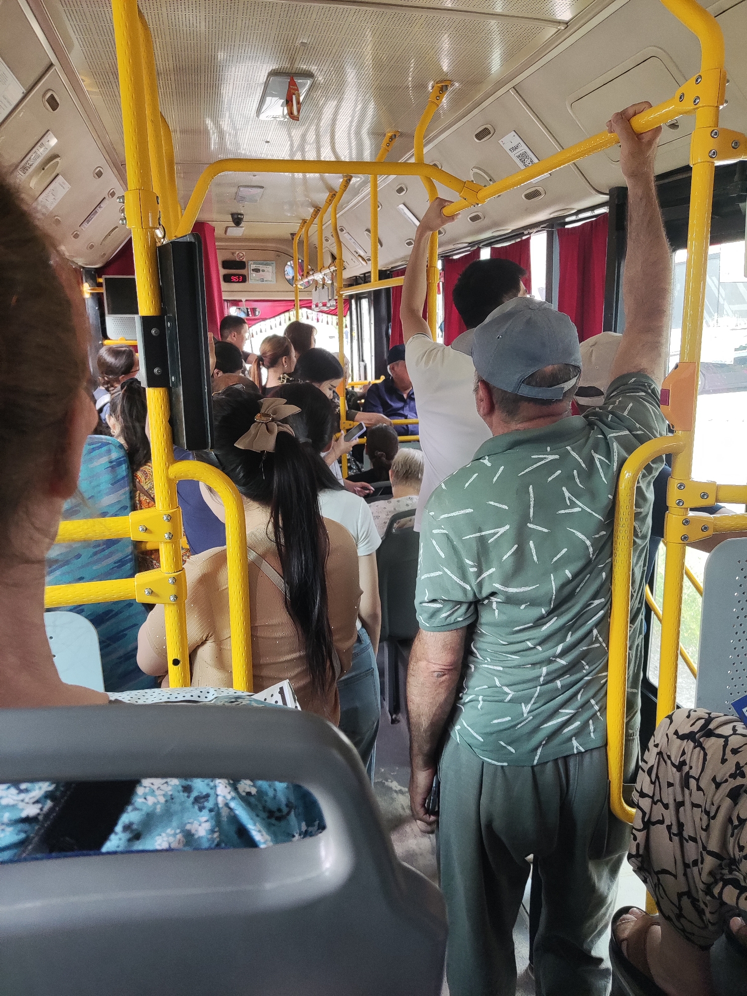 An interior view of a crowded public bus shows passengers standing and holding onto yellow handrails. The bus is brightly lit, with red curtains visible in the background. A digital display above the passengers shows "9:53". The scene appears to be in Shymkent, Kazakhstan.