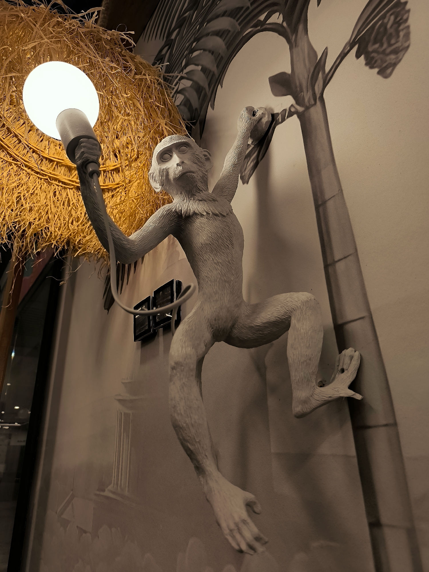 An artistic sculpture of a monkey is mounted on a wall, mimicking it climbing a tree. The monkey is depicted in a dynamic pose, with one hand holding a light bulb that illuminates a nearby straw-like lamp shade. The background features a mural or wallpaper with a jungle theme, including palm fronds and architectural elements. Two black electrical outlets are visible on the wall below the monkey. The lighting in the scene is dim, with the primary light source being the bulb held by the monkey. This artistic installation is located in the city administration building of Qarağandı, Kazakhstan.