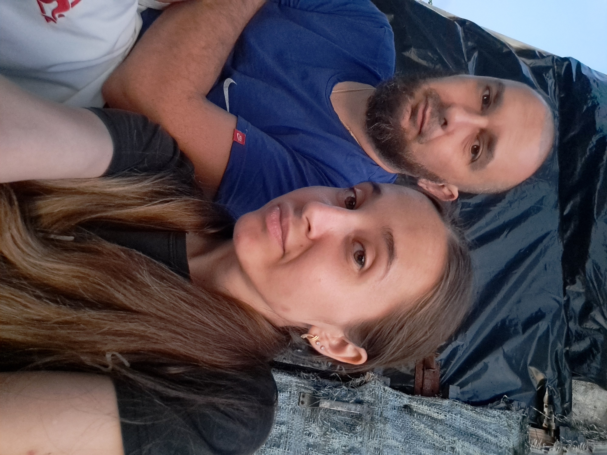 The image is a close-up, outdoor selfie of a man and a woman sitting together. The man, on the left, has a shaved head, a beard, and is wearing a blue t-shirt with a Nike logo on the sleeve. He is looking directly at the camera with a neutral expression. The woman, on the right, has long, brown hair and is wearing a dark t-shirt. She is also looking at the camera with a slight smile and is wearing earrings. They appear to be in a rustic or outdoor setting, with a dark tarp and textured material visible in the background. The sky is visible at the top left, suggesting daytime. The presence of a blue tarp, possibly used for shelter or protection, suggests a location that might be undergoing construction, renovation, or is a temporary dwelling. The overall impression is a casual, intimate moment captured between two individuals. The image does not provide specific geographical markers to confirm its location in Vypasne, Ukraine.