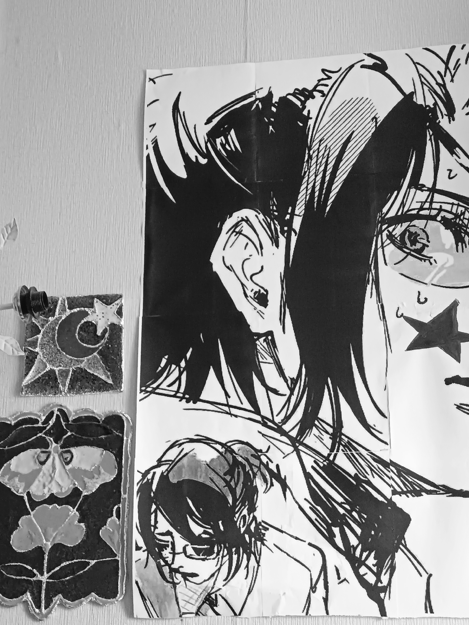 The image shows a section of a wall in what appears to be an indoor space. The wall is textured. Hanging on the wall is a large, black-and-white drawing of what seems to be a manga-style character. The drawing occupies a significant portion of the frame. To the left of the drawing are two smaller, square-shaped decorations or artwork. The top one features a crescent moon and star motif, while the bottom one depicts a flower. There are also elements of a hanging light fixture in the upper left corner. There is no definitive information to say with certainty that the image was taken in the city of Посад, Russia.