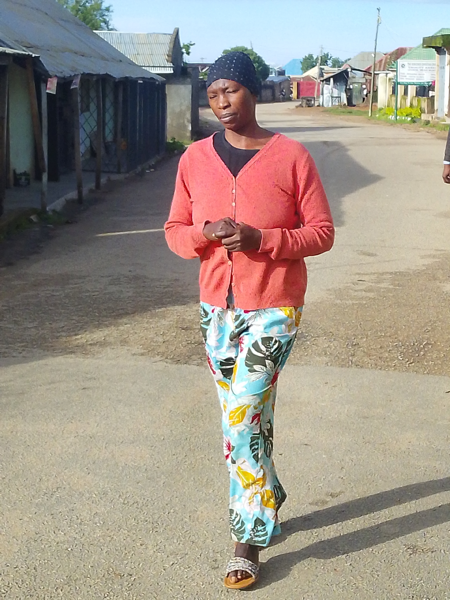 A woman walks down a paved street lined with shops and buildings. She is wearing a black head covering, a coral pink cardigan over a black shirt, and brightly colored floral patterned pants. Her hands are clasped in front of her. The street appears to be in a town or city, with corrugated metal roofs visible on some of the buildings. The lighting suggests it is daytime, possibly late afternoon due to the shadows cast on the street. There are no other people clearly visible in the immediate vicinity. The overall impression is of a residential or commercial street in a developing urban area.