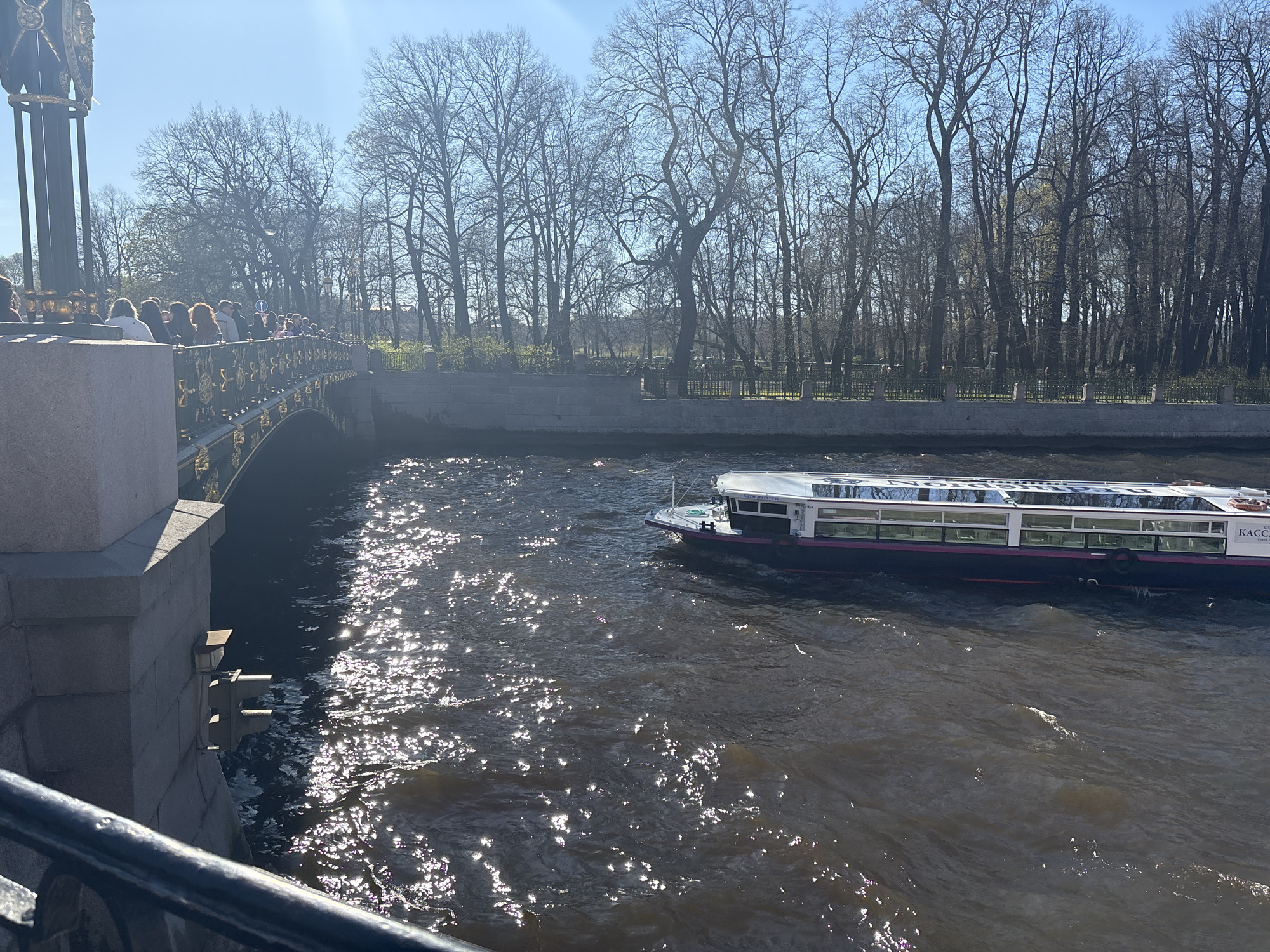 A low-angle, eye-level shot captures a canal scene in St. Petersburg, Russia, on a sunny day. The waterway dominates the frame, its surface reflecting sunlight. A boat is navigating to the right of the frame. A bridge, adorned with decorative metalwork and people crossing it, spans the canal to the left. Bare trees line the canal banks in the background.
