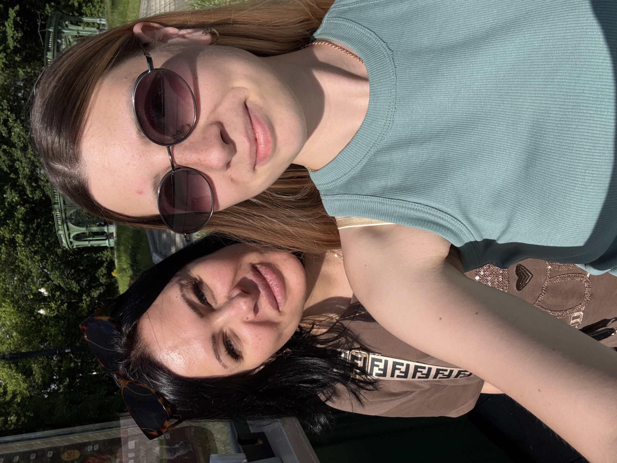 This media file captures a bright outdoor selfie of two women, likely taken in Moscow, Russia. Both women are smiling subtly, suggesting a pleasant and relaxed mood.
The woman positioned higher in the frame has long, light brown hair and wears round, dark-tinted sunglasses. A delicate gold chain necklace is visible around her neck, and she is dressed in a light blue ribbed tank top. The strong sunlight illuminates her face, highlighting her features.
The woman positioned lower in the frame has dark hair and a small nose piercing. Her tortoiseshell-patterned sunglasses are pushed up onto her head. She wears a brown top, and a prominent strap with the repeated "FENDI" logo in a light color is visible across her chest, possibly part of a bag or garment.
The background features lush green foliage, indicating an outdoor setting such as a park or garden. A light-colored, ornate structure, possibly a gazebo or pavilion with columns, is partially visible through the trees, suggesting a well-maintained public space. The clear, sunny weather is evident from the bright lighting and distinct shadows cast across the subjects and environment, pointing to either morning or afternoon. The overall impression is one of leisure and enjoyment on a sunny day.