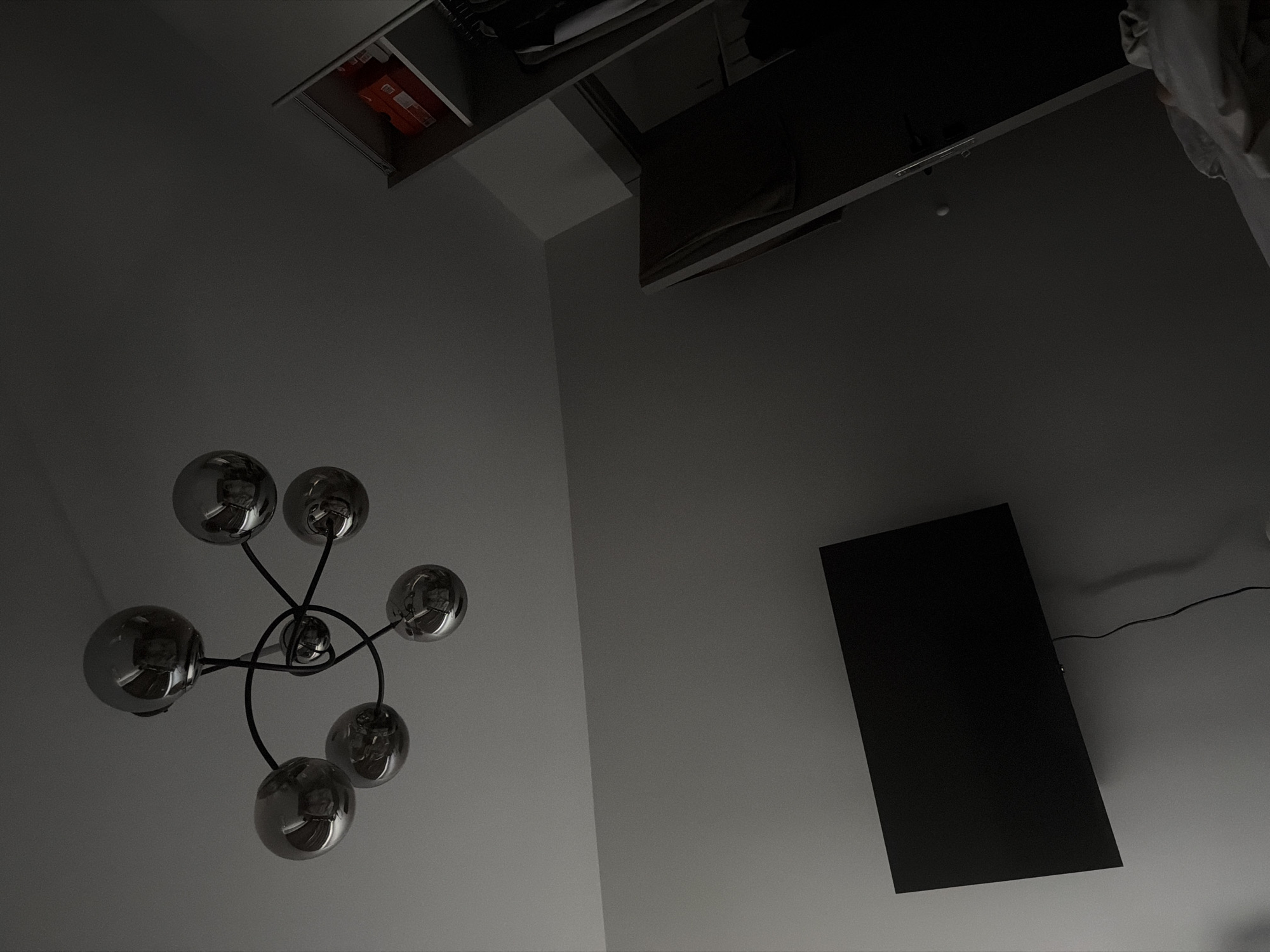 The image presents a low-angle shot of an interior space with modern elements. A multi-bulb chandelier with reflective, spherical shades is visible on the left side of the frame against a light-colored wall. In the upper portion of the image, a shelving unit or cabinet appears to be mounted near the ceiling. A flat-screen television is positioned on the right side of the frame, with its screen facing the viewer. A power cable extends from the television toward the right edge of the image. The room's lighting is subdued.
There is no indication of the image being taken in Plovdiv, Bulgaria or any other location.