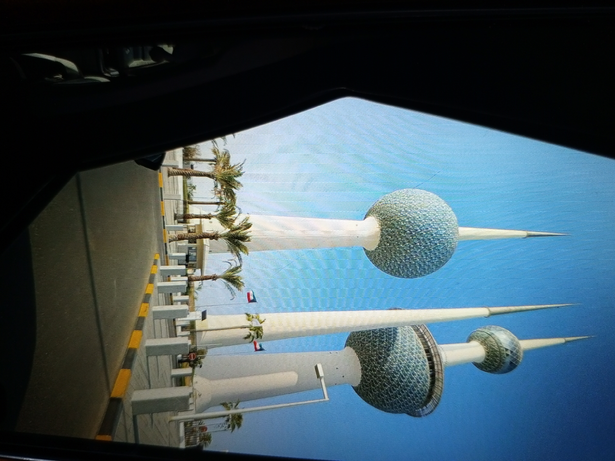 The image shows a side view of the Kuwait Towers, which are a group of three stainless steel towers in Kuwait City, Kuwait. The towers are located on an inlet of the Persian Gulf. The image is taken from a slightly elevated position, possibly from a vehicle, looking towards the towers. The foreground features a road with yellow and black striped barriers and palm trees lining the side. The towers are tall and slender, with spherical upper sections covered in a mosaic of blue and green tiles. They are topped with pointed spires. The sky is a clear blue. The reflection of the towers is visible on a surface, possibly water or a screen, creating a mirrored effect. The image appears to be a photograph or screenshot, possibly of a digital display, as indicated by the presence of what seems like a screen edge at the top and bottom of the frame and a slight pixelation or screen effect. The location of Golbembal, Nigeria is not represented in this image.