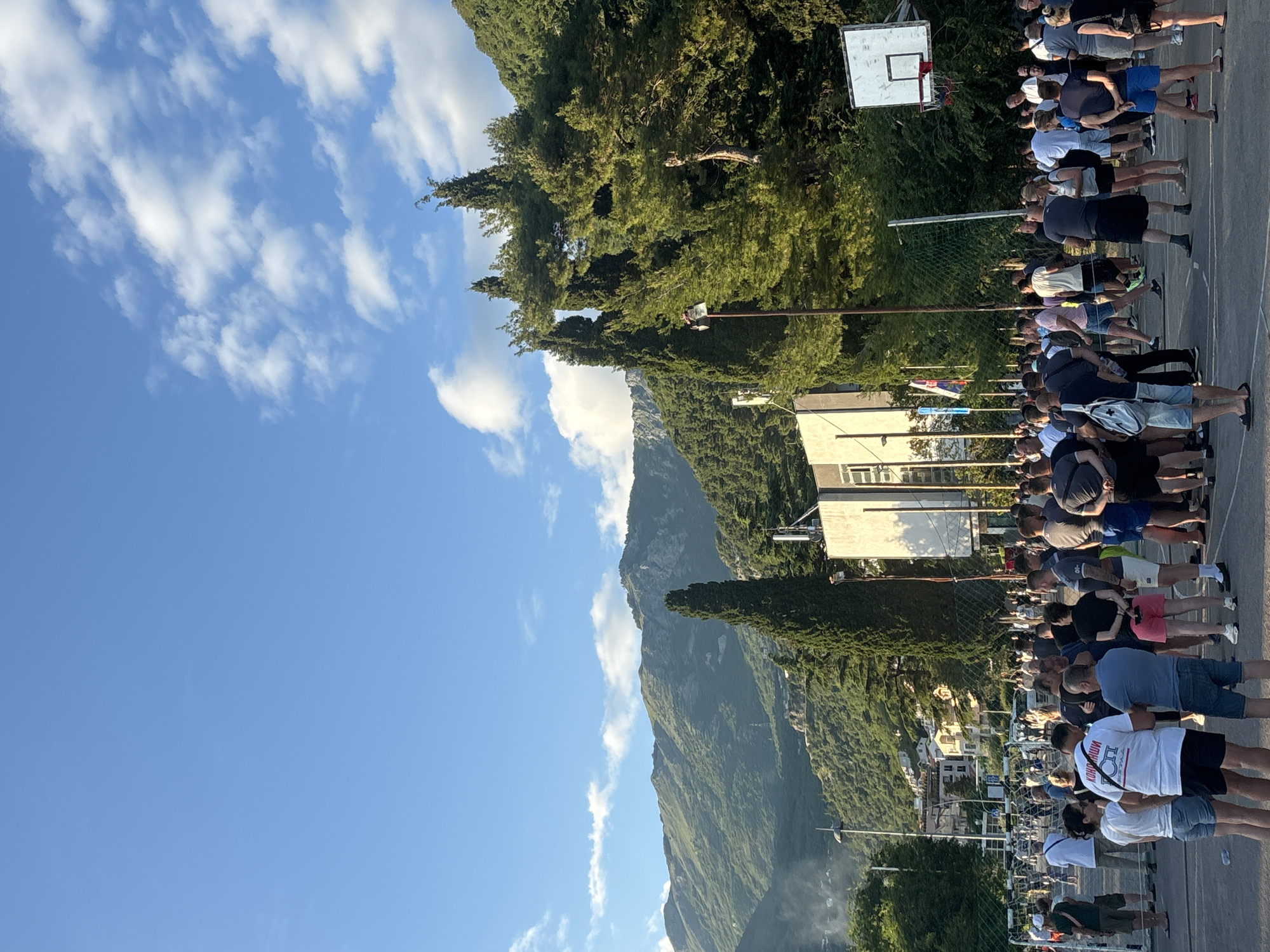 The image depicts an outdoor gathering of numerous people in Budva, Montenegro, under a clear blue sky dotted with scattered white clouds. The scene is brightly lit by daylight.
In the foreground and mid-ground, a large group of individuals, predominantly adults and appearing to be mostly men, are standing in several organized rows on a paved surface, likely a sports court. They are dressed in casual summer wear, including shorts and t-shirts, and most face away from the camera. A basketball hoop with a white backboard and a net are visible to the right, reinforcing the sports court setting.
The background is dominated by a majestic, verdant mountain range on the left, with its steep slopes densely covered in green trees and foliage. To the right of the mountains, several light-colored buildings are visible, suggesting urban or residential structures. More trees are interspersed throughout the landscape.
The people appear to be observing an event or waiting in line. Visible text on a white t-shirt worn by a person in the center-left reads "КОНВОЙ" (Konvoy). Another person on the far right has partially visible text that appears to be "YUGOSLAVIA" or similar. The overall mood seems calm and orderly within this scenic mountainous environment.