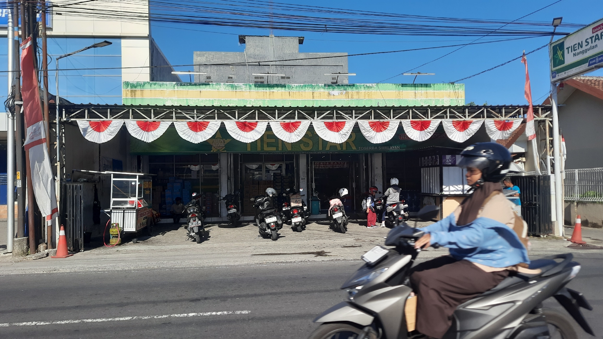 In Nanggulan, Indonesia, a street scene unfolds under a clear blue sky. A building with a green awning and white and red decorative fans spans the width of the frame, displaying the sign "TIEN STAR TOSERBA SWALAYAN." In front of the building, several motorcycles are parked. To the left, a food cart with a gas cylinder sits by a fence. In the foreground, a person on a motorcycle, wearing a black helmet and a blue jacket over a brown garment, is captured in motion, facing right. A smaller flag on a pole is visible to the right of the building, accompanied by a green sign that reads "TIEN STAR Nanggulan." The street itself is paved, with white lines marking the lanes.
