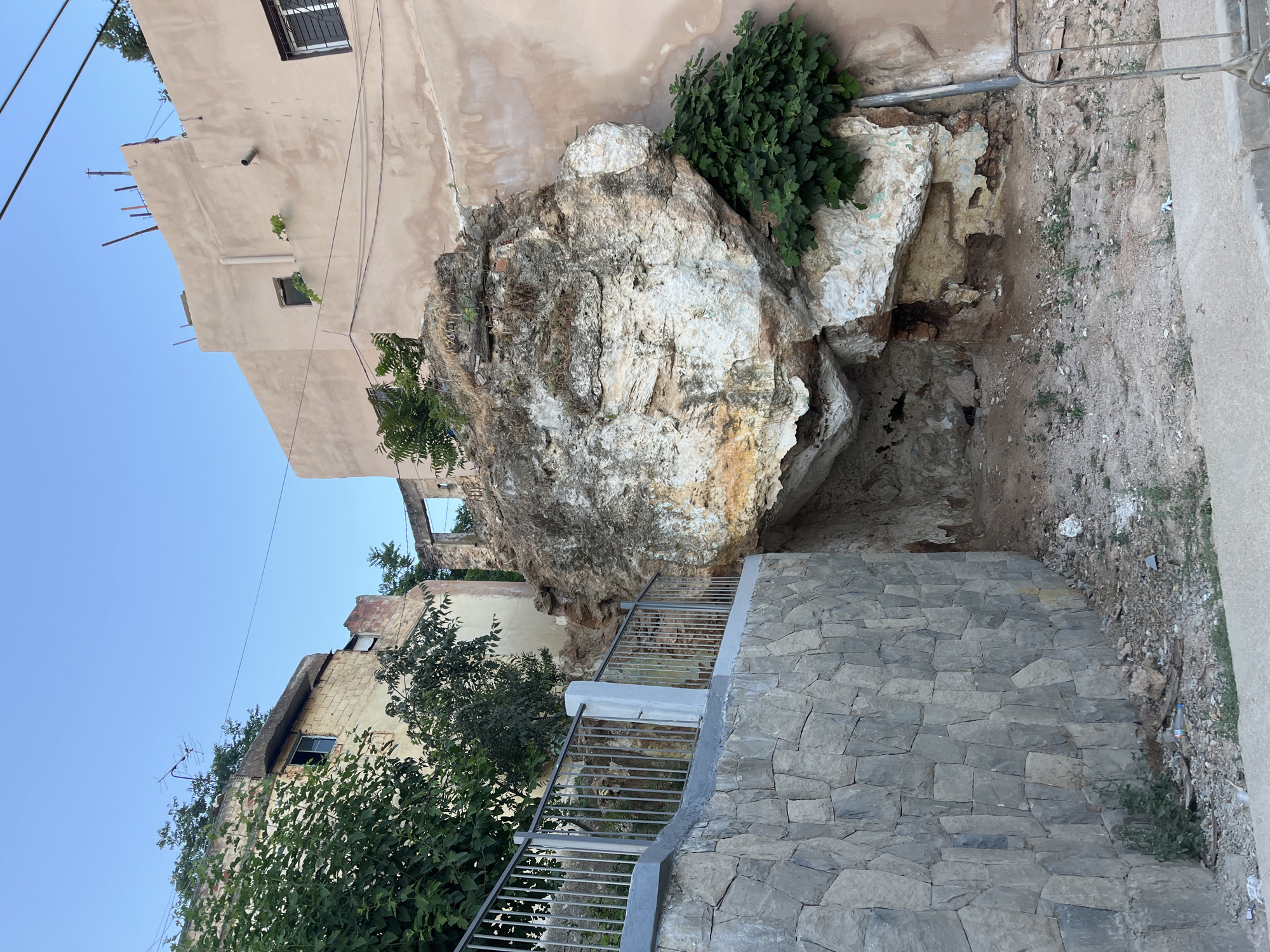 The image depicts an urban scene in El Hajeb, Morocco, characterized by a large, weathered rock formation that juts out from a building's foundation. The rock, a mix of light and dark brown with white and greenish patches, appears to have weathered and eroded over time, revealing an internal cave-like structure at its base. Green vegetation, possibly fig trees, grows around the rock and on top of the adjacent building.
To the left of the rock formation, a constructed stone wall with a metal railing runs along a raised pathway. The wall is made of irregularly shaped gray stones. Behind the wall, trees and parts of buildings are visible. The buildings themselves appear to be old and in a state of disrepair, with exposed brickwork, broken windows, and peeling paint. The architecture suggests a historical or traditional Moroccan style.
In the foreground, the ground is covered with dirt, gravel, and scattered debris, including a discarded plastic bottle. The overall environment appears somewhat neglected, with evidence of both natural elements (the rock, vegetation) and human habitation (buildings, walls, debris). The sky above is clear and blue, indicating daytime. There are no people visible in the image. The scene conveys a sense of age and the passage of time, with natural forces and urban decay coexisting.