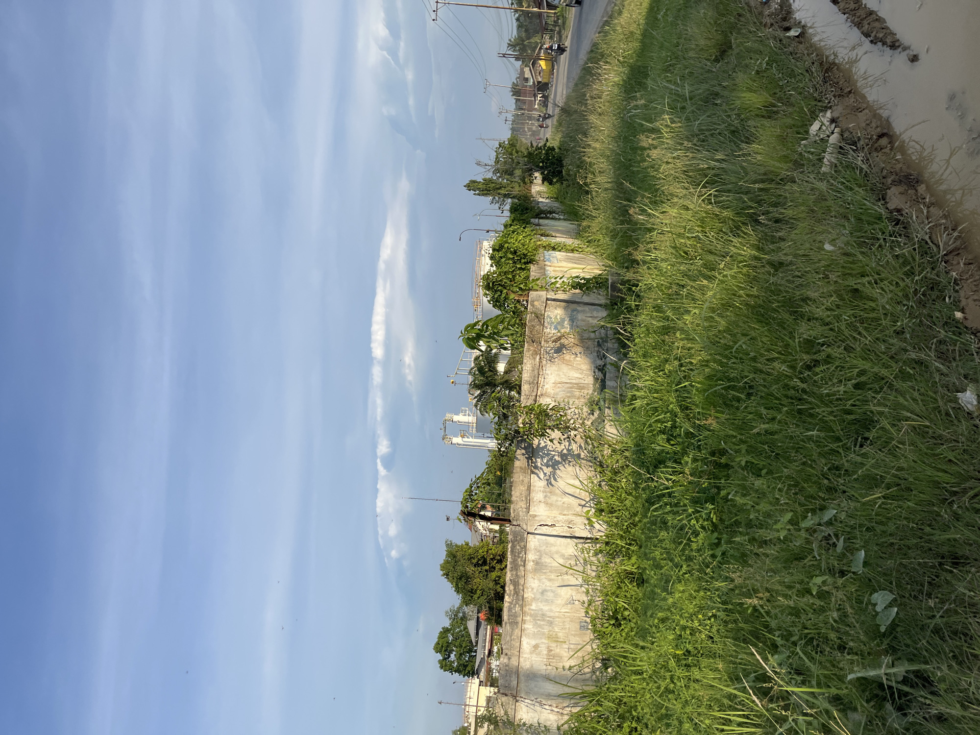 The image depicts an outdoor scene in Alurcucur, Indonesia, under a partly cloudy sky. In the foreground, tall green grass grows along a dirt path that leads to a muddy area with tire tracks. A low, cracked concrete wall runs horizontally across the middle ground, separating the grassy area from a road. Beyond the wall, buildings and trees are visible, including industrial structures with white tanks and pipes. The road to the right is lined with utility poles and power lines. Several motorcycles and a car are present on the road, with a few distant figures. Birds are scattered in the sky. The overall impression is one of an undeveloped or industrial fringe area within the city.