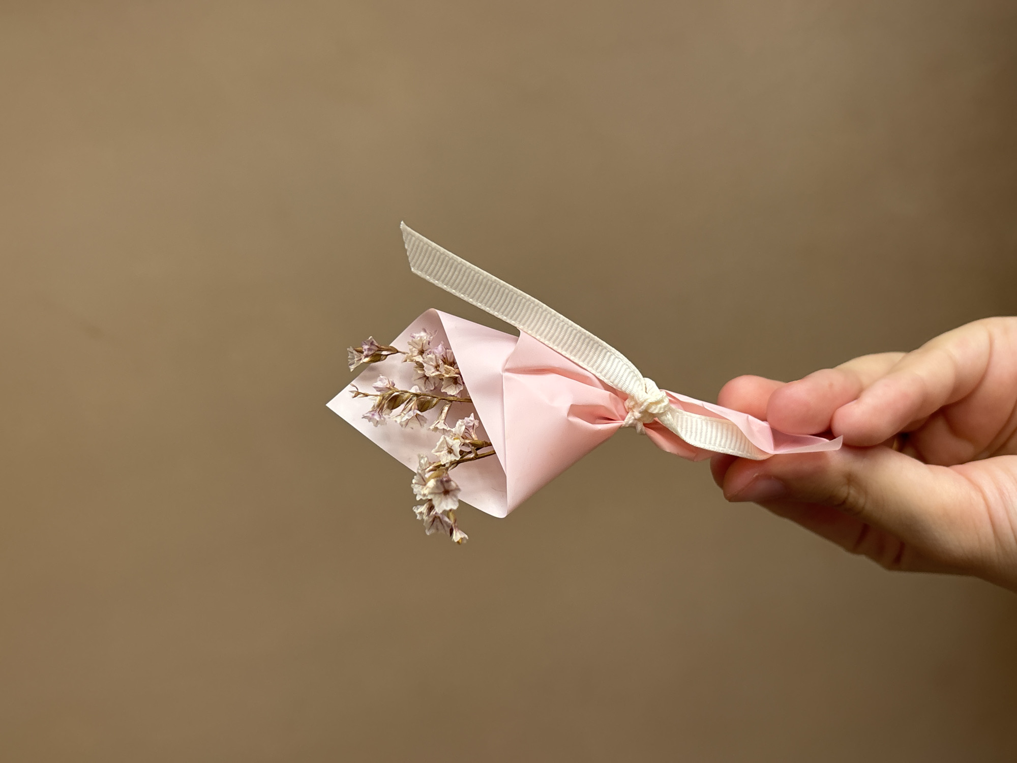 A hand holds a small bouquet of dried flowers wrapped in pink paper and tied with a cream-colored ribbon. The bouquet is held up against a plain, muted brown background. The focus is on the delicate flowers and their packaging, with the hand only partially visible, suggesting a close-up shot. The lighting is soft, creating a gentle atmosphere. No specific time of day or weather conditions are evident. The setting appears to be indoors with a simple, neutral backdrop. The image conveys a sense of simplicity and natural beauty. There is no visible text in the image. The location context of Orihuela, Spain, is not visually represented in the image itself.