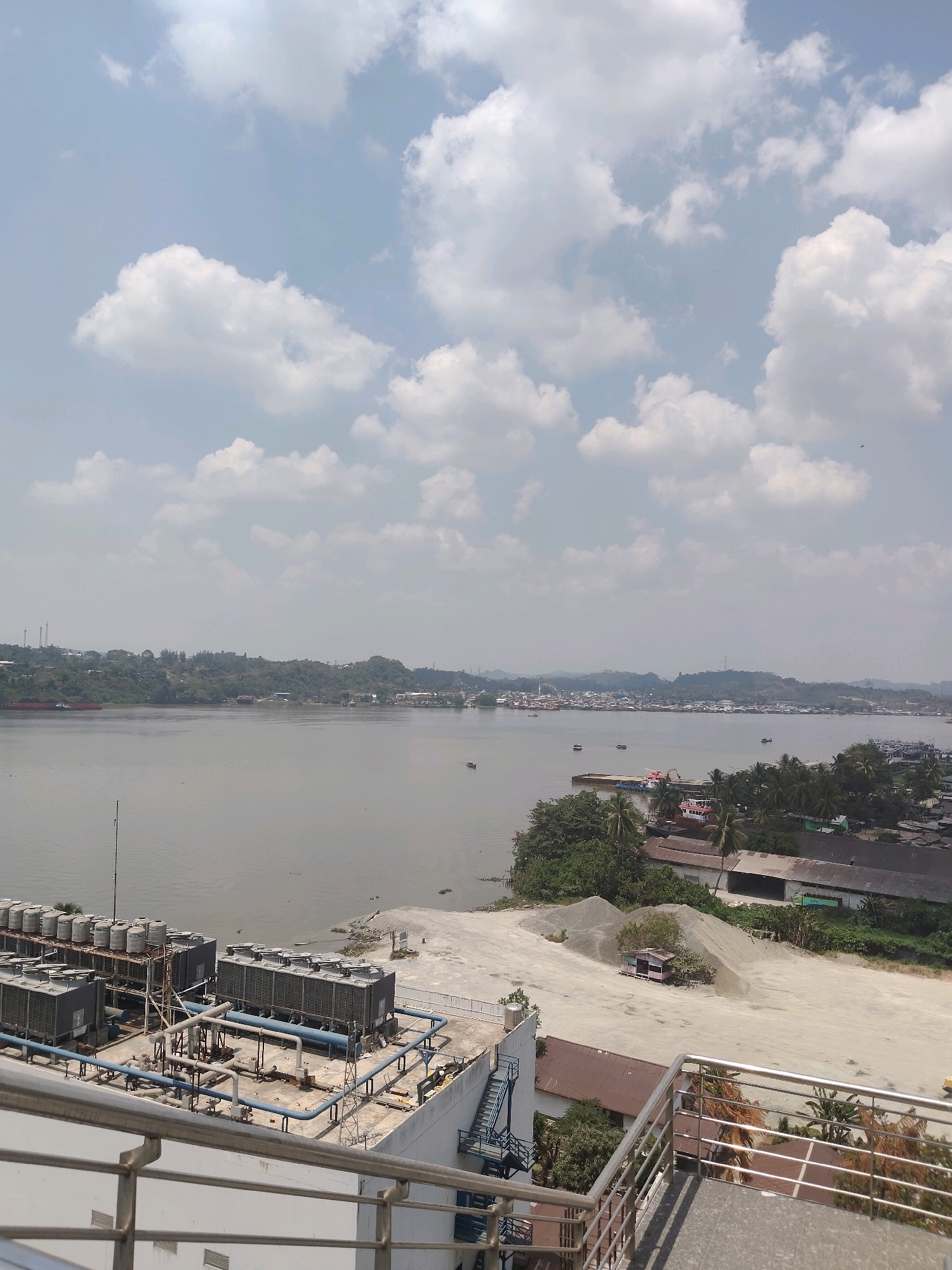 The image shows a wide river with a distant shoreline dotted with buildings and lush green hills under a partly cloudy sky. In the foreground, a rooftop is visible, featuring several large air conditioning units and a complex network of pipes. The river itself appears calm, with a few small boats and barges scattered across its surface. Along the shoreline, a town or city is visible, with structures ranging from modest houses to larger industrial buildings. The overall impression is one of a semi-urban environment situated alongside a significant body of water. This scene is consistent with views along the Mahakam River in Samarinda, Indonesia, a city known for its extensive river system and maritime activity.