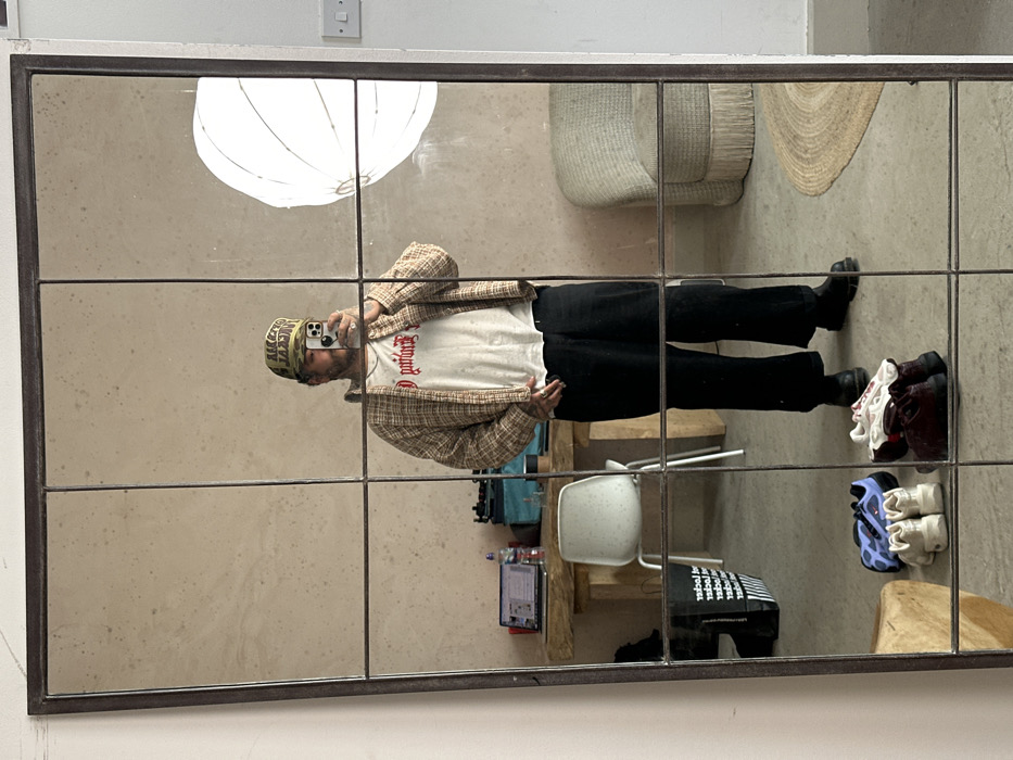 A full-length, multi-paneled mirror reflects a male individual taking a selfie with a smartphone in an interior space in London, United Kingdom. The individual is centrally positioned, wearing a camouflage baseball cap, a white t-shirt with red gothic-style text, an open beige and red plaid shirt, dark wide-leg trousers, and dark boots. Tattoos are visible on the hands and forearms, and rings are worn on the fingers. The smartphone obscures the individual's upper face, revealing a beard.
Reflected on the left side of the image, behind the individual, is a rustic wooden table with an open laptop, a water bottle, and other miscellaneous items. A white plastic chair is situated behind the table. A black tote bag with white text, including "LEATHER LANE," rests on the floor to the left of the table.
On the right side of the image, a large white spherical light diffuser or softbox is visible, emitting light. Below it, a light-colored, textured armchair with a fringed base sits on a circular woven rug. A white wall with a standard UK double light switch is also visible.
The floor is a light grey concrete or similar material, and the walls are a textured, light beige. In the foreground reflection, several pairs of shoes are arranged on the floor, including blue and black sneakers, white sneakers, and dark red/burgundy shoes. The "LEATHER LANE" text on the tote bag indicates the location is associated with or in the vicinity of Leather Lane, Holborn, within the City of London.