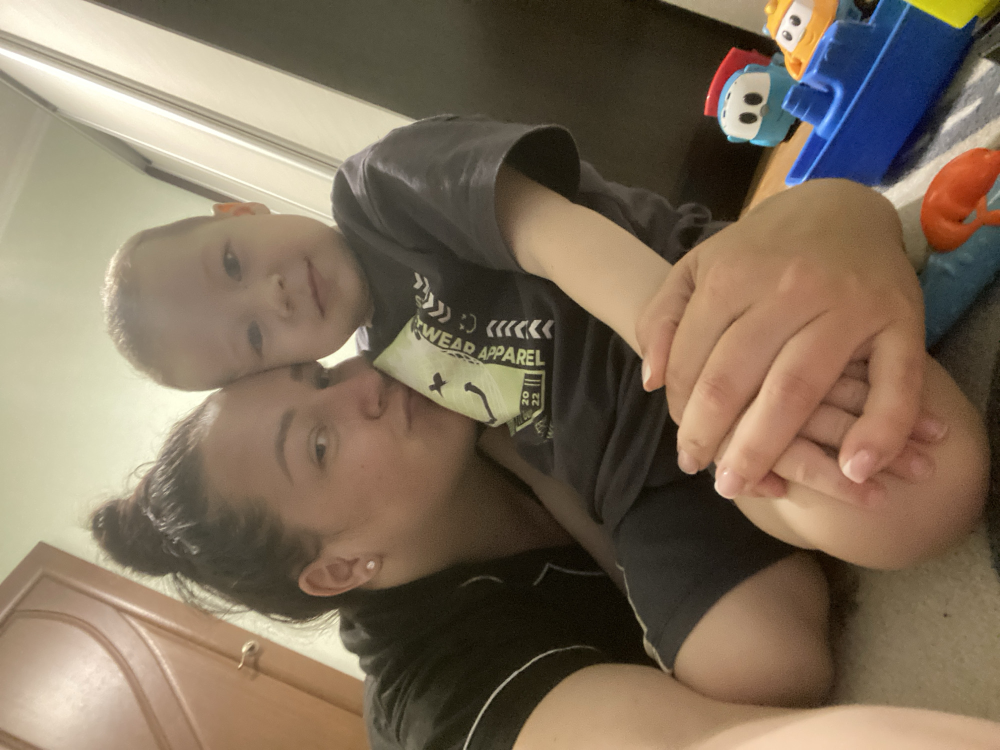 A young child is lying on top of an adult, possibly a parent, in what appears to be an indoor setting. The child is wearing a dark-colored t-shirt with a light-colored graphic design. The adult is wearing a dark top. A collection of toys is visible to the right of the subjects. A door can be seen in the bottom left corner of the image. The photograph appears to have been taken from a low angle. There is no evidence of the location being Sochi, Russia.