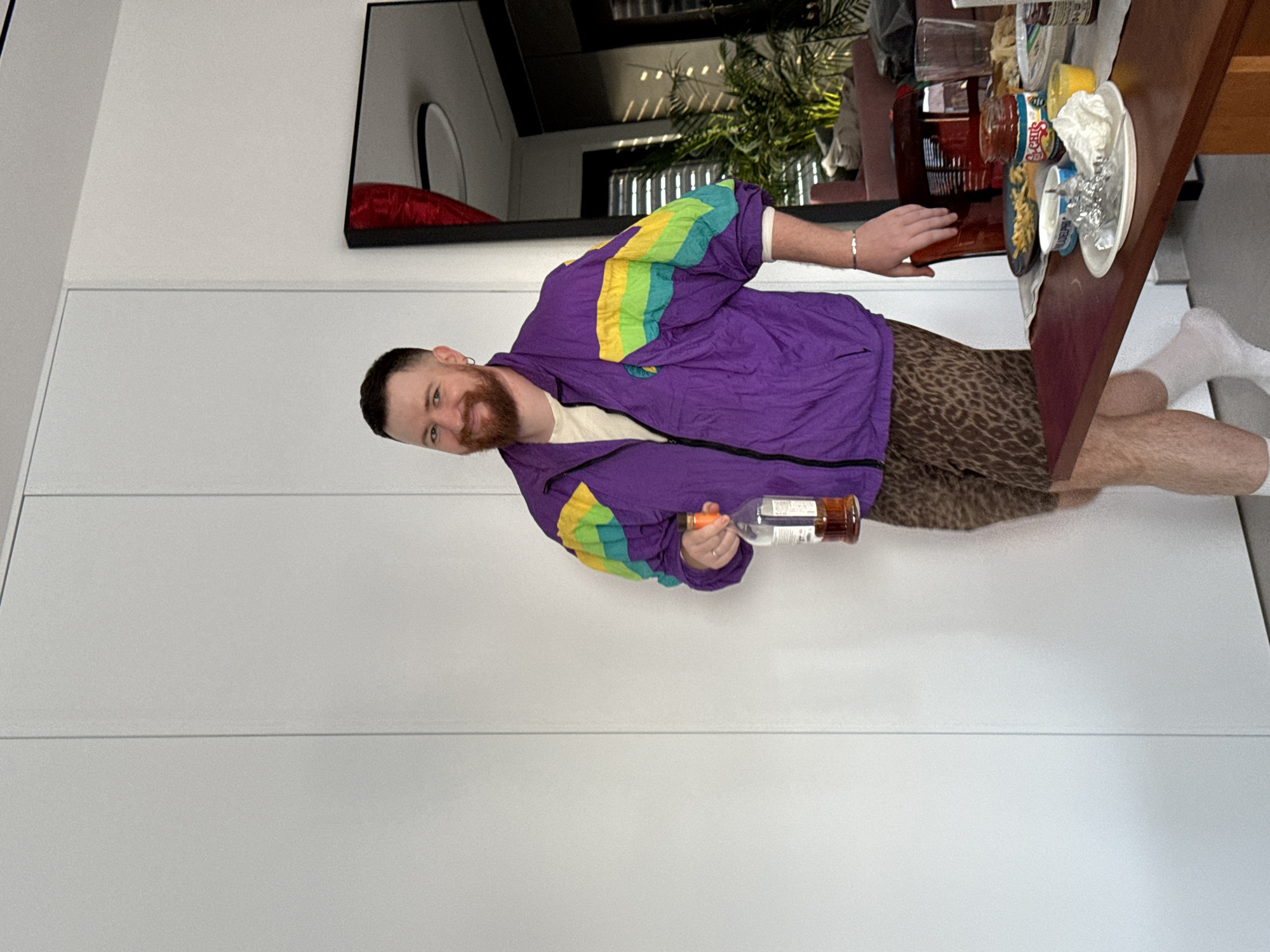 A person stands indoors next to a table cluttered with various items, including food, jars, and other containers. The person is wearing a purple jacket with colorful shoulder accents and leopard-print shorts. They are holding a bottle in one hand. The background features a white wall with a modern black-framed mirror and possibly a closet or storage space. The location in the city of Tel-Aviv, Israel is not discernible from the image alone.