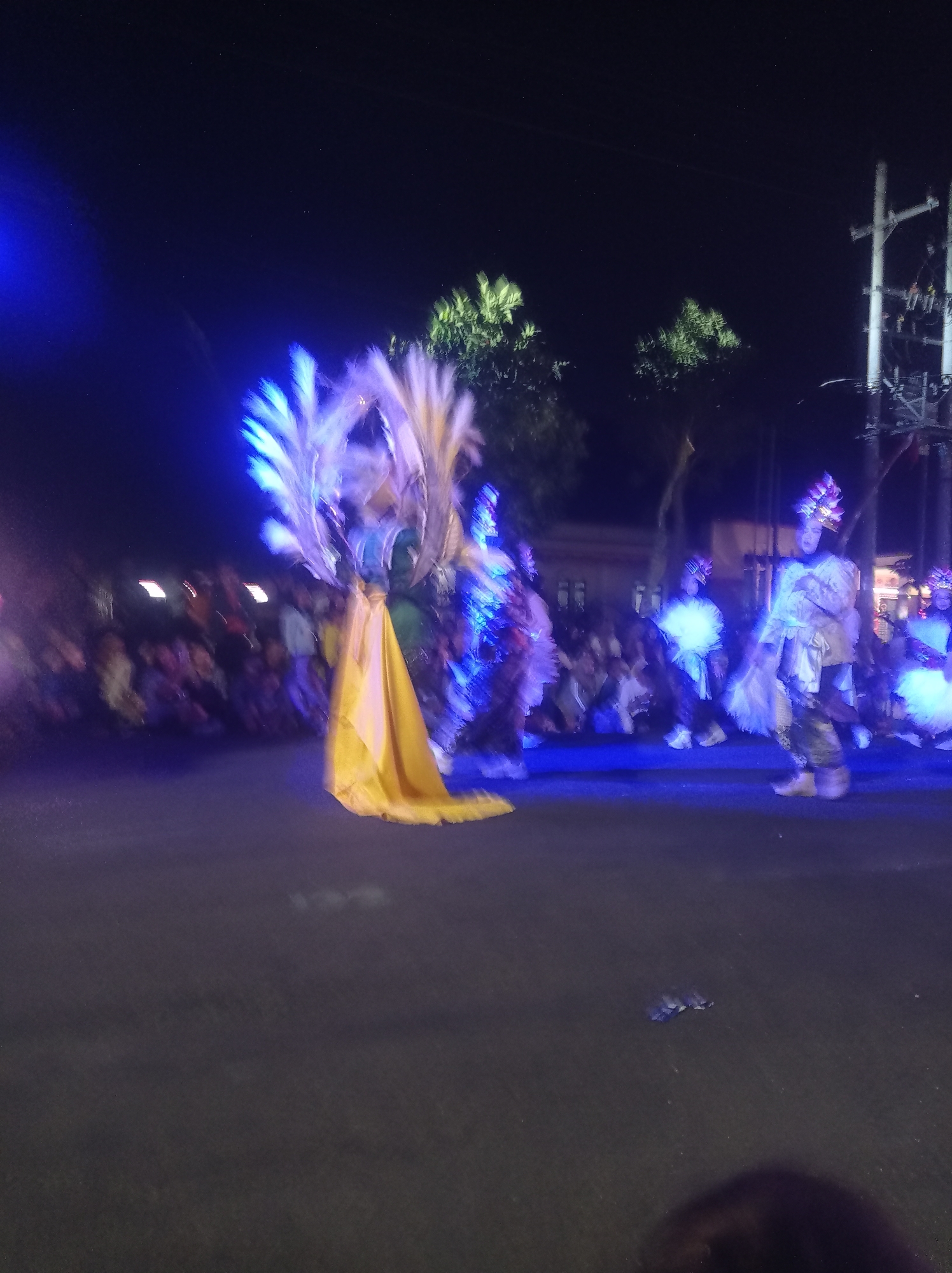 This image captures a nighttime street performance in Pandean, Indonesia. A performer in a vibrant yellow costume adorned with large, feathered headdresses is the central focus, illuminated by bright blue lights. Several other performers in elaborate, illuminated costumes are visible to the right, also participating in the event. A crowd of spectators is present, lining the street and observing the parade. The background reveals trees and a utility pole, indicating an urban outdoor setting. The scene appears to be part of a festival or celebration, with a lively atmosphere suggested by the movement and light.