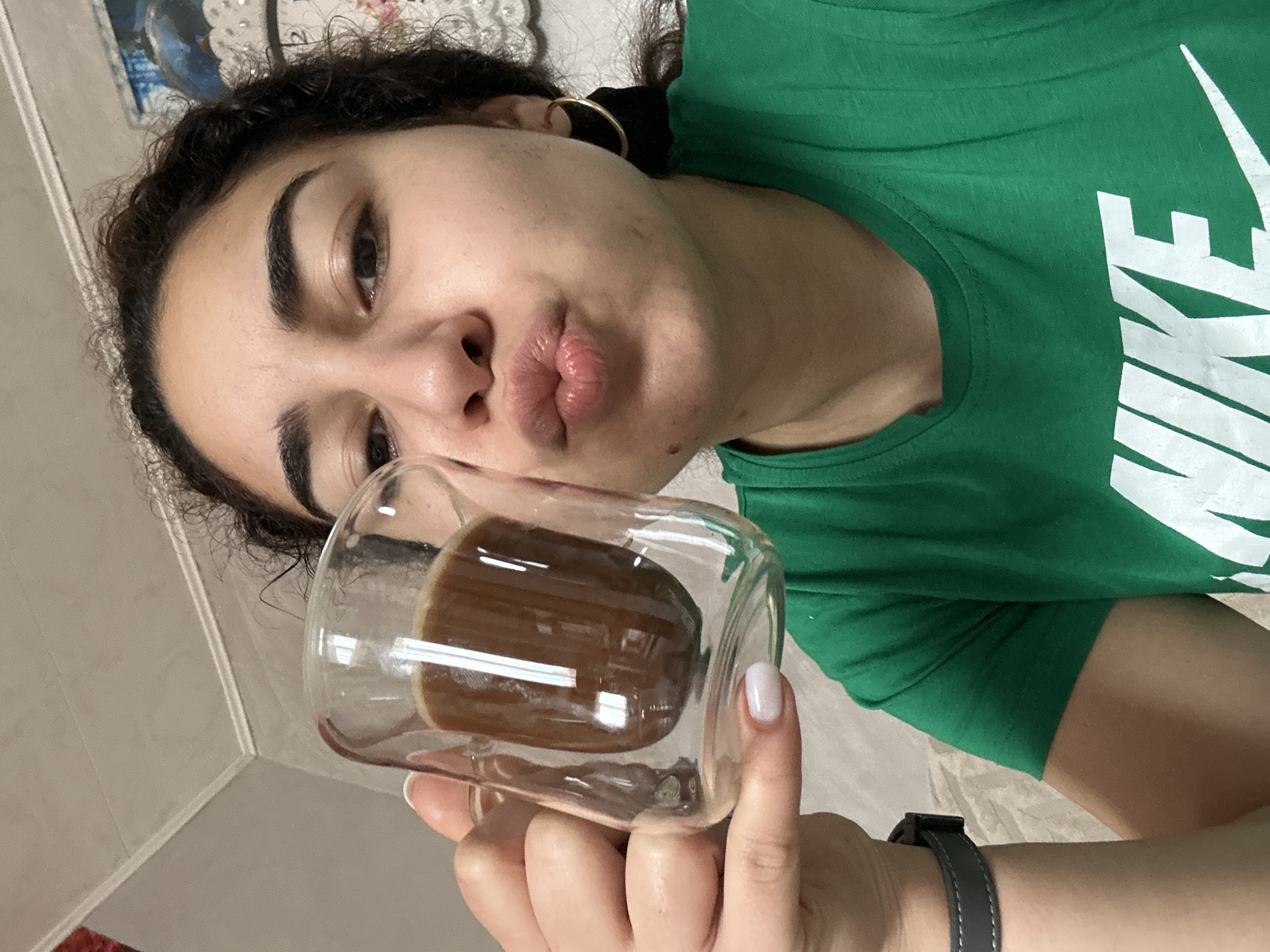 A woman is holding a glass of coffee in front of her face. She is wearing a green Nike T-shirt and a watch. She has curly dark hair and is wearing a gold hoop earring. She is puckering her lips. The background is a neutral color. The image does not contain any information suggesting Burgas, Bulgaria or any other location.