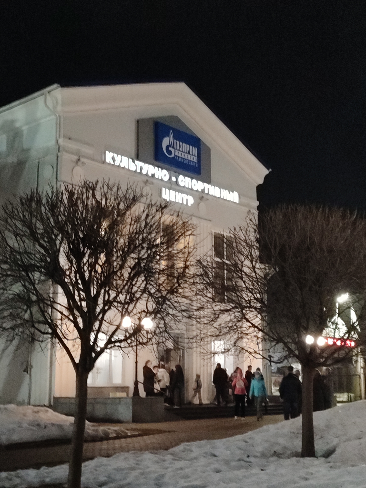 The photo shows the Gazprom Cultural and Sports Center in Chaikovsky, Russia, at night. The building is white with a classical facade. A blue sign with the Gazprom logo is visible above a neon sign in Russian. Several people are gathered on the steps leading up to the building. The ground is covered in snow, and bare trees are visible in the foreground. Streetlights illuminate the scene.