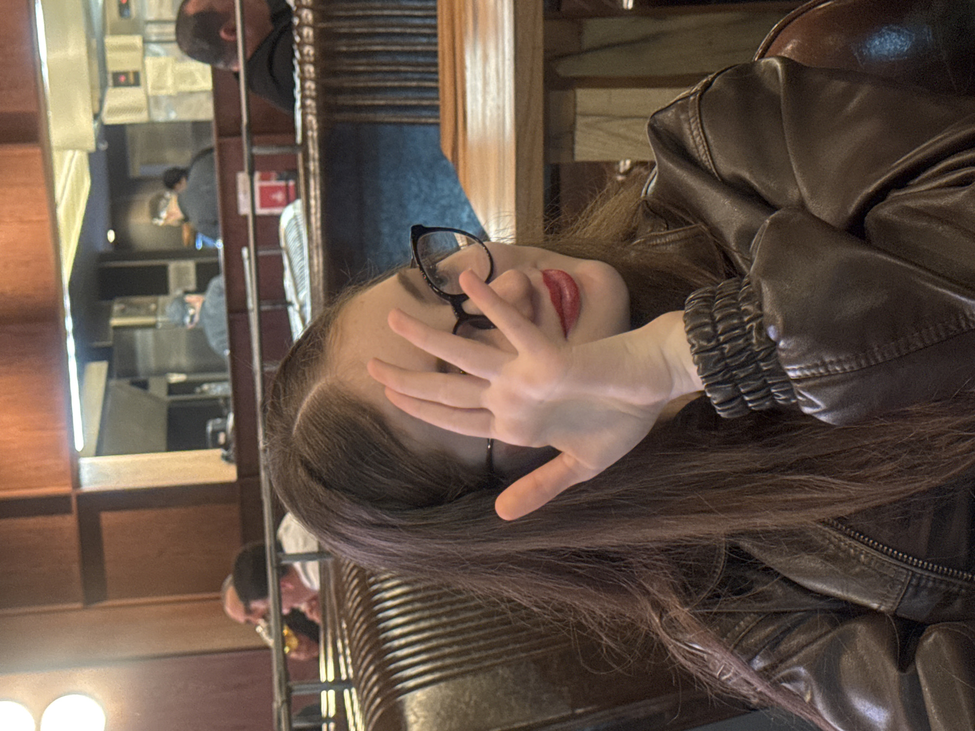 A woman with long brown hair and glasses wears a brown leather jacket while seated at a restaurant. She holds her hand up towards the camera as if to block the view. The restaurant appears to be in Tbilisi, Georgia. In the background, a reflection shows staff working in the kitchen, and other patrons are seated at tables.