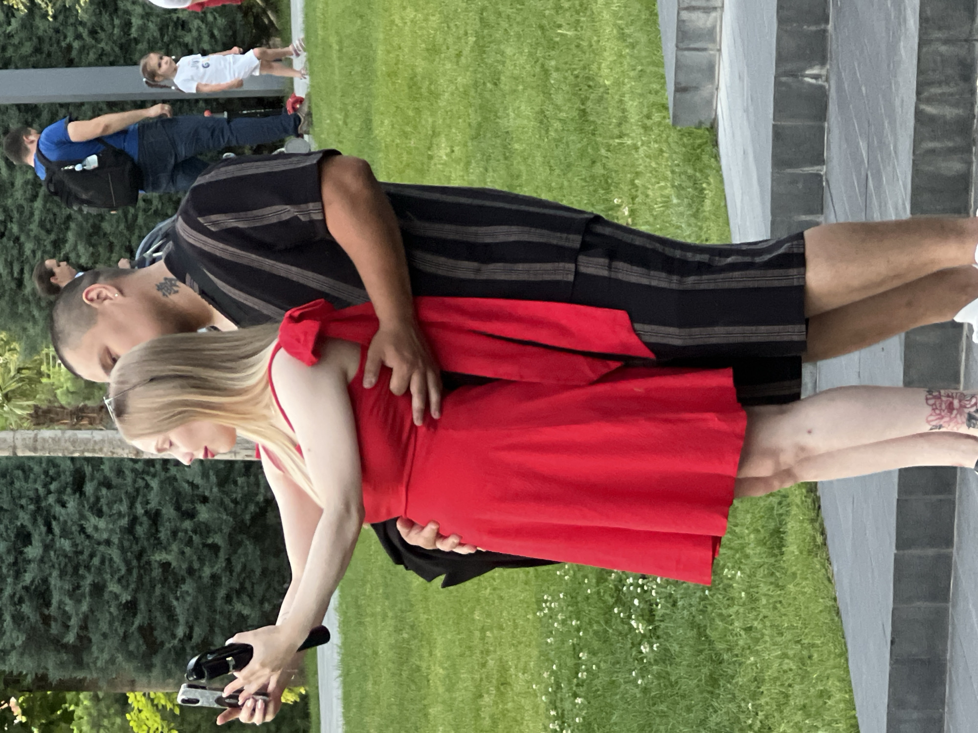 A man and a woman are posing for a selfie on stone steps in a park. The woman, in a red dress, is holding a smartphone and a selfie stick. The man, in a black and white striped shirt and shorts, has his arm around her. In the background, other people are walking and a child is standing. The scene is set in a city park in Krasnodar, Russia, with lush green trees and grass.