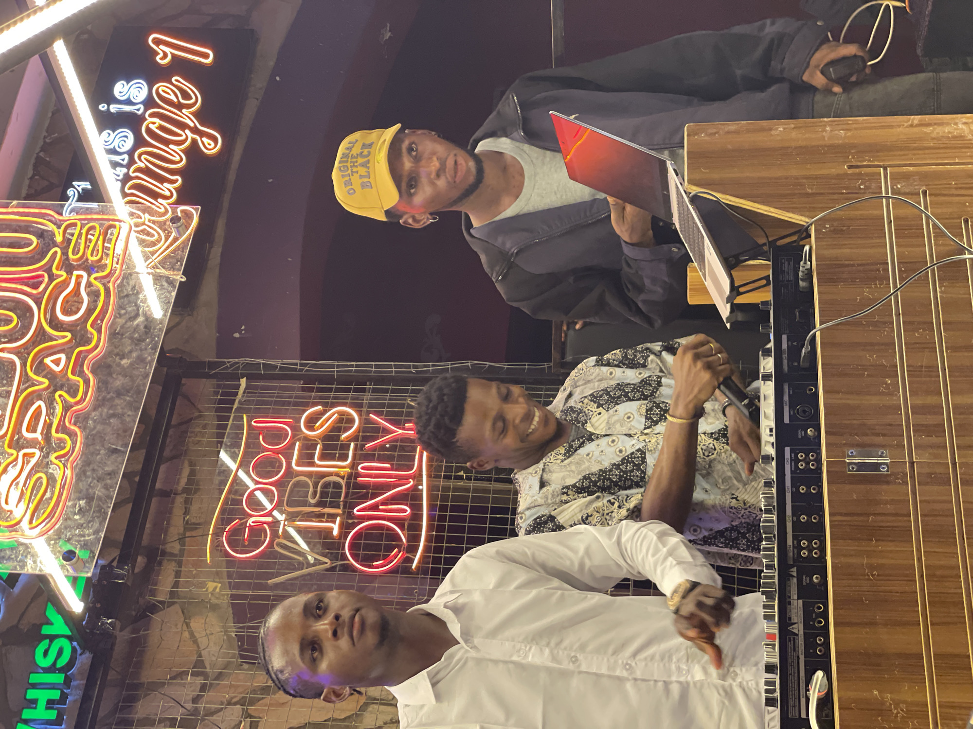 Three men are gathered around a sound mixing board. The man at the top wears a yellow hat and a jacket over a t-shirt, and is working on a laptop. The man in the middle wears a patterned shirt and holds a microphone. The man in the front wears a white shirt. Behind them is a neon sign that reads "Good Vibes Only," and another sign with “Lounge 1." The location is likely in Lokoja, Nigeria, given the implied context.